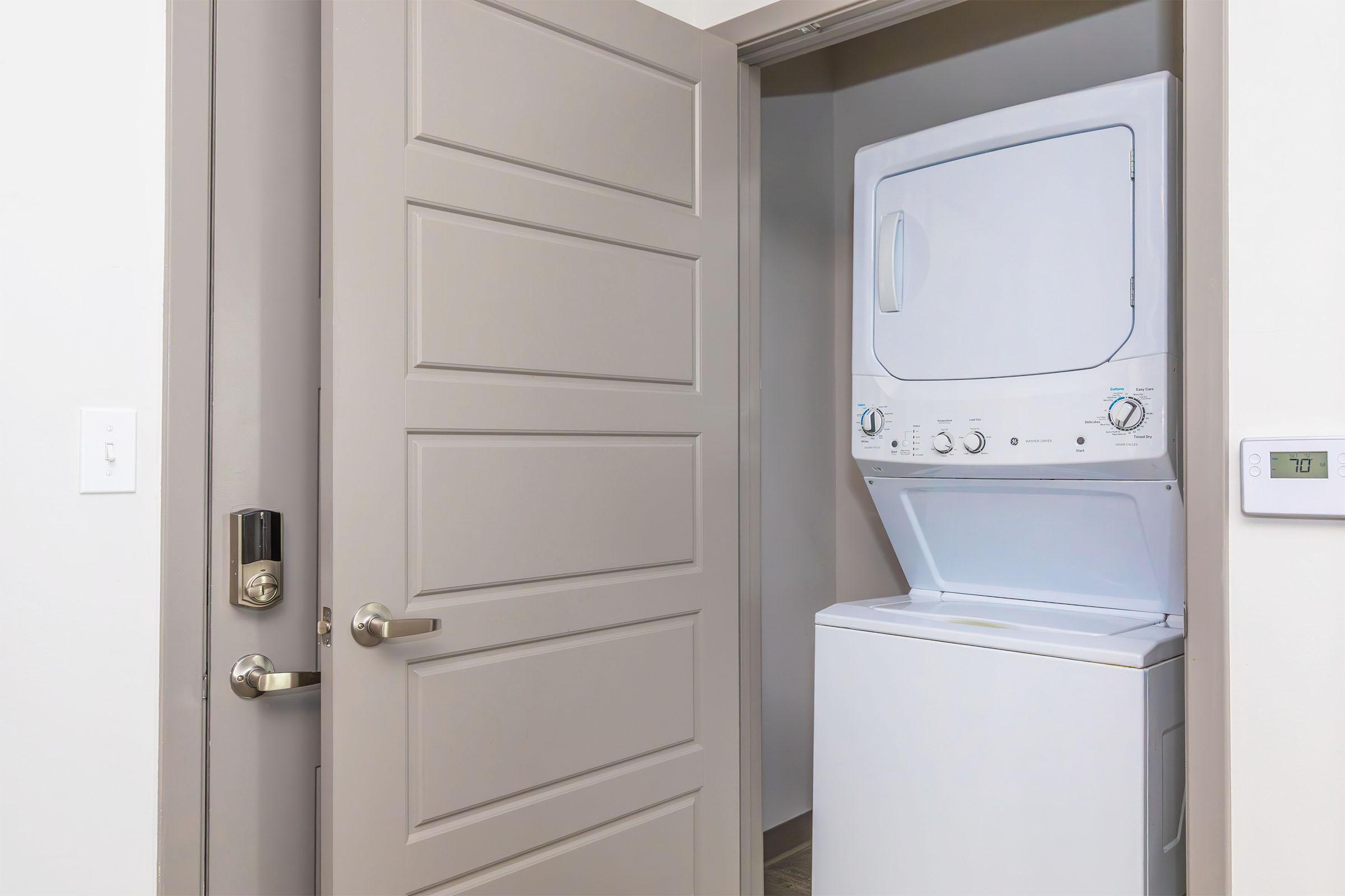 Stacked washer and dryer located in a small laundry area, visible through an open door. The door is a light color with a modern design, and a digital thermostat is mounted on the wall beside the door. The overall space appears clean and well-lit.