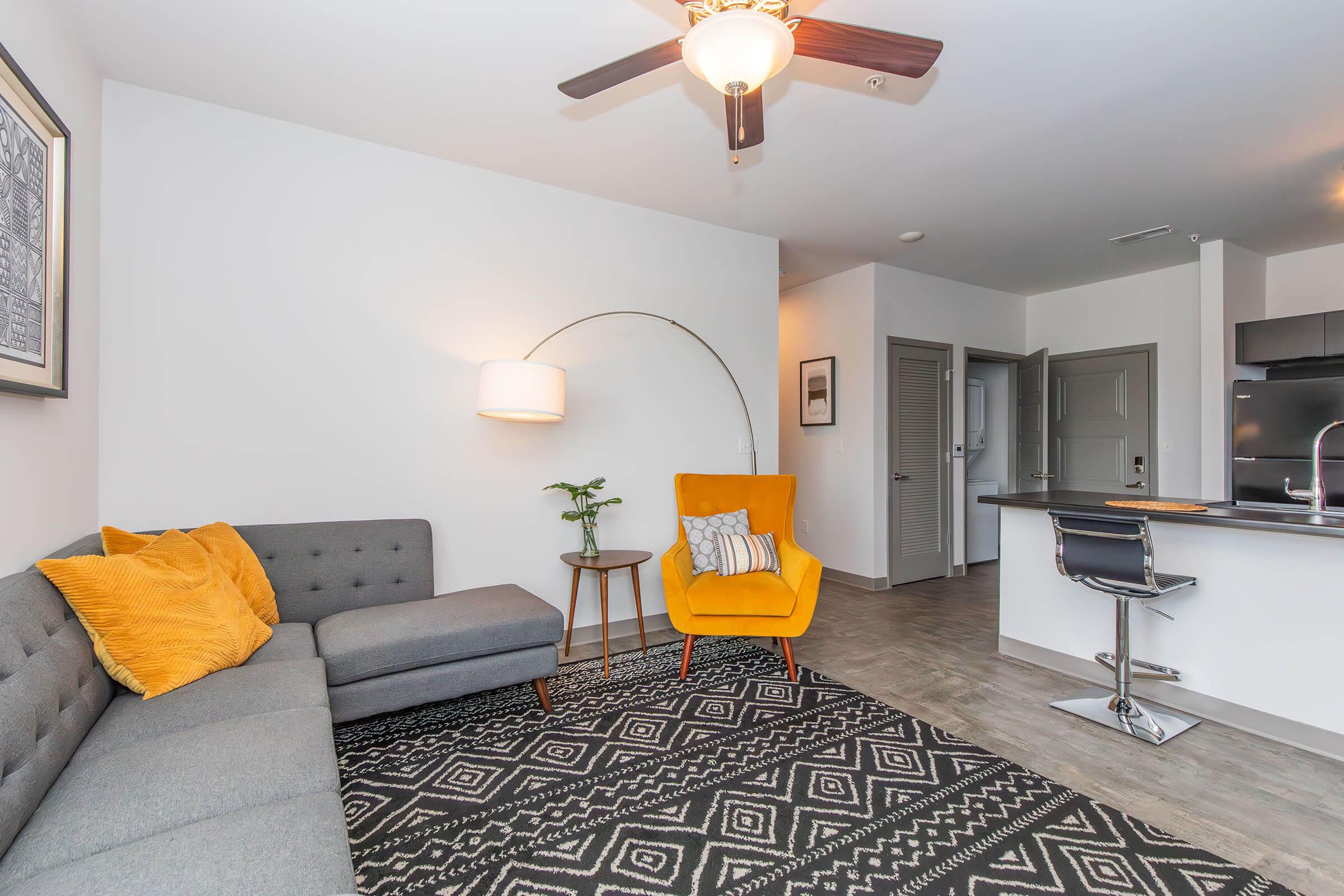 A modern living room featuring a gray sectional sofa with orange throw pillows, a bright yellow accent chair, and a stylish arc floor lamp. There is a small side table with a plant, and a kitchen area is visible in the background with a bar counter and stools. The flooring is a light wood finish.