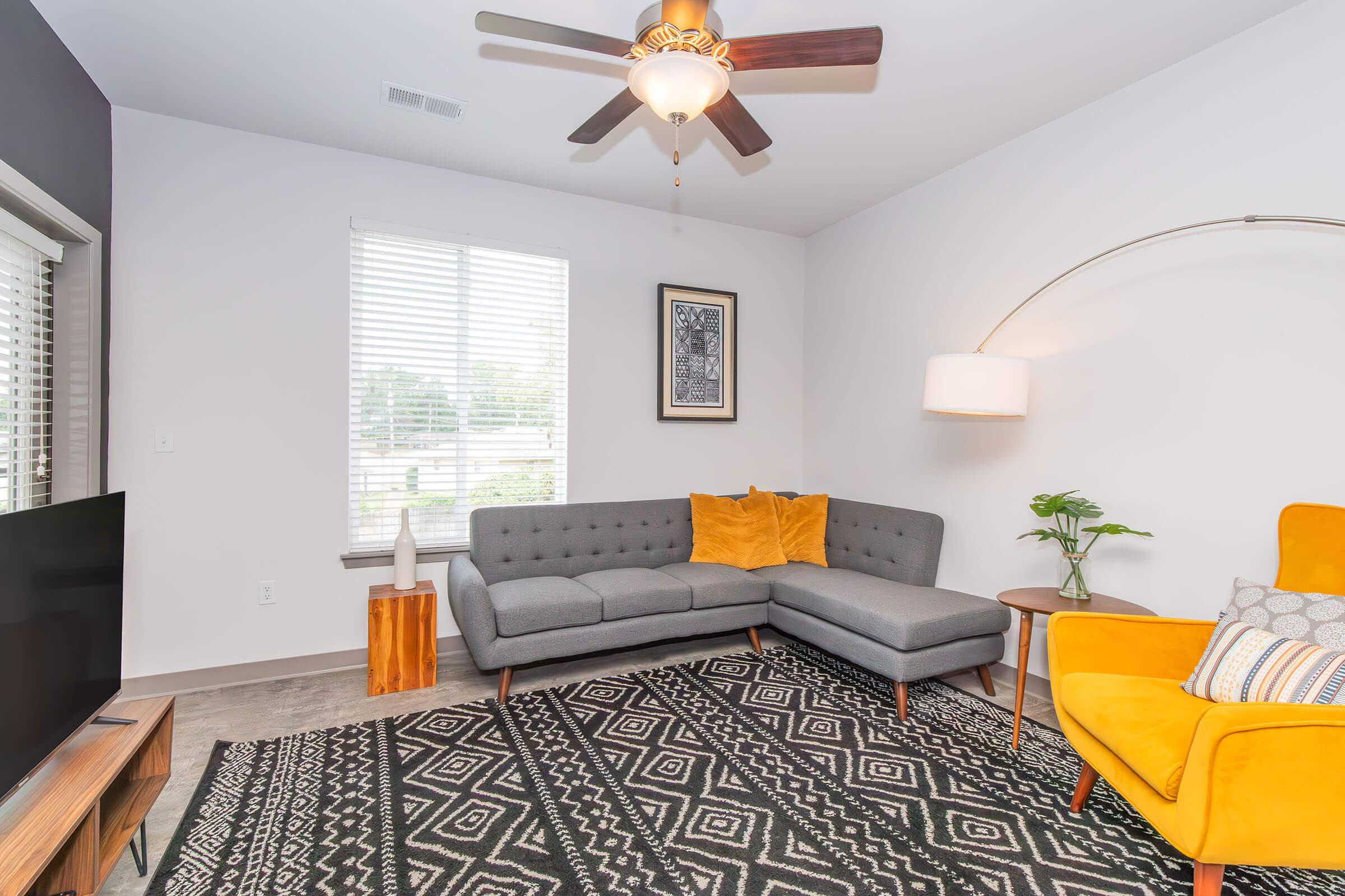 Stylish modern living room featuring a gray sectional sofa with yellow throw pillows, a yellow accent chair, a round side table, and a ceiling fan. A large TV sits on a wooden media console. Decorative plants and an abstract wall art piece enhance the cozy atmosphere, complemented by natural light from a window with blinds.