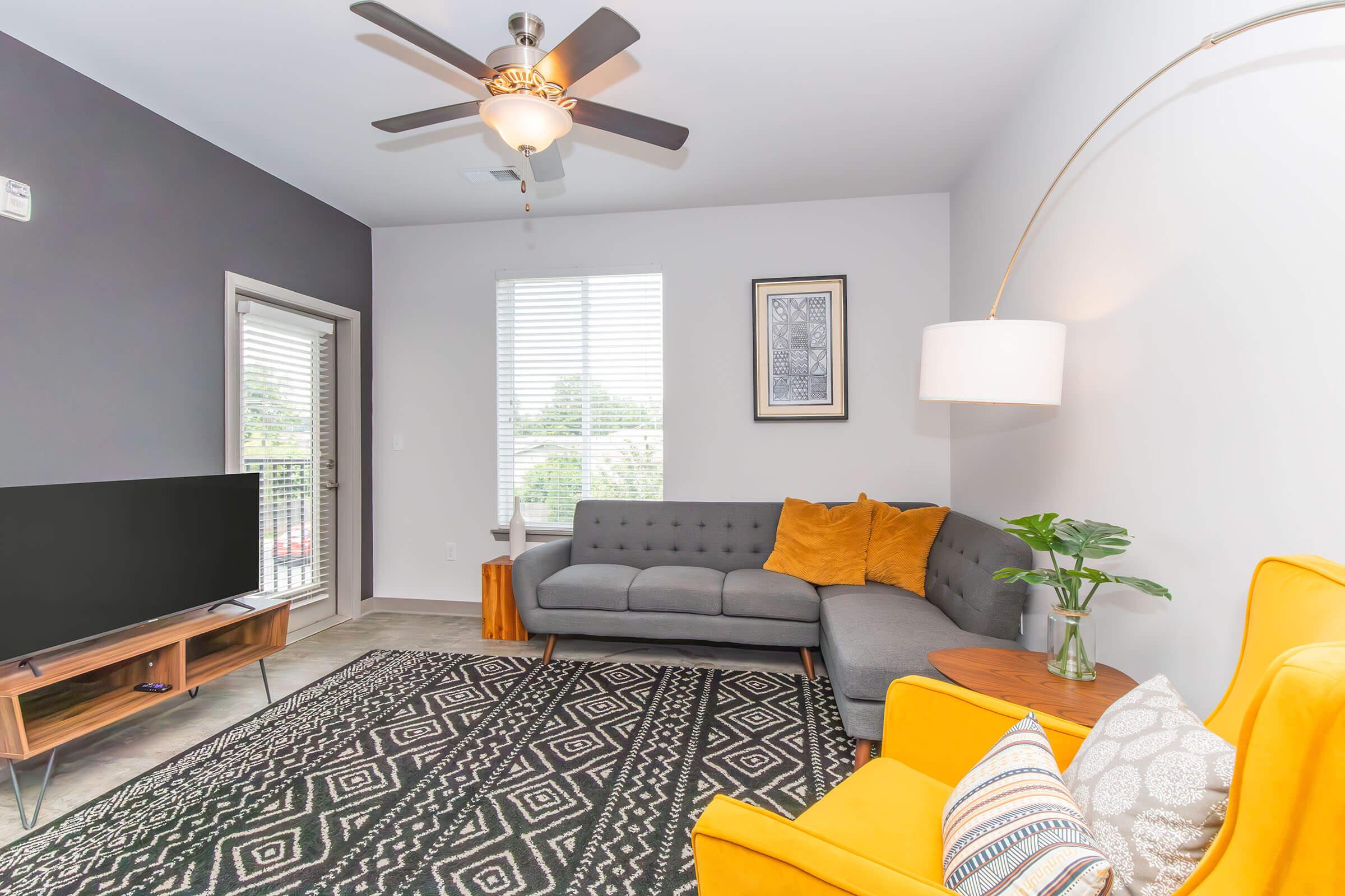 A modern living room featuring a gray sofa with orange cushions, a yellow accent chair, and a wooden television stand with a TV. A large window allows natural light in, and a stylish area rug lies on the floor. A ceiling fan and arc floor lamp add contemporary decor elements.