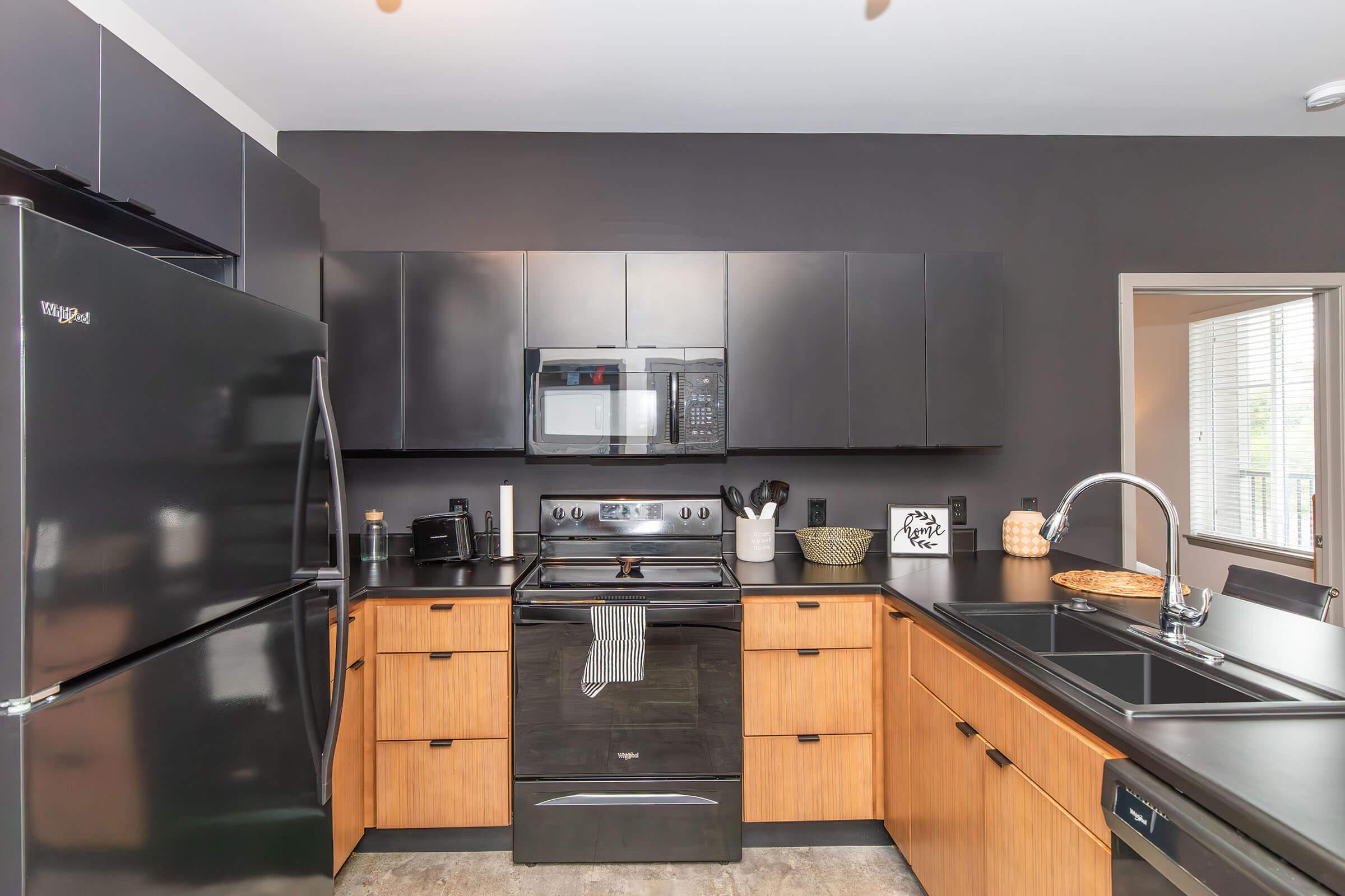Modern kitchen featuring dark gray walls, black appliances, and wooden cabinetry. The layout includes a black stove and microwave, a large black sink, and a stylish countertop. There are decorative elements like a bowl and artwork, alongside a window providing natural light.