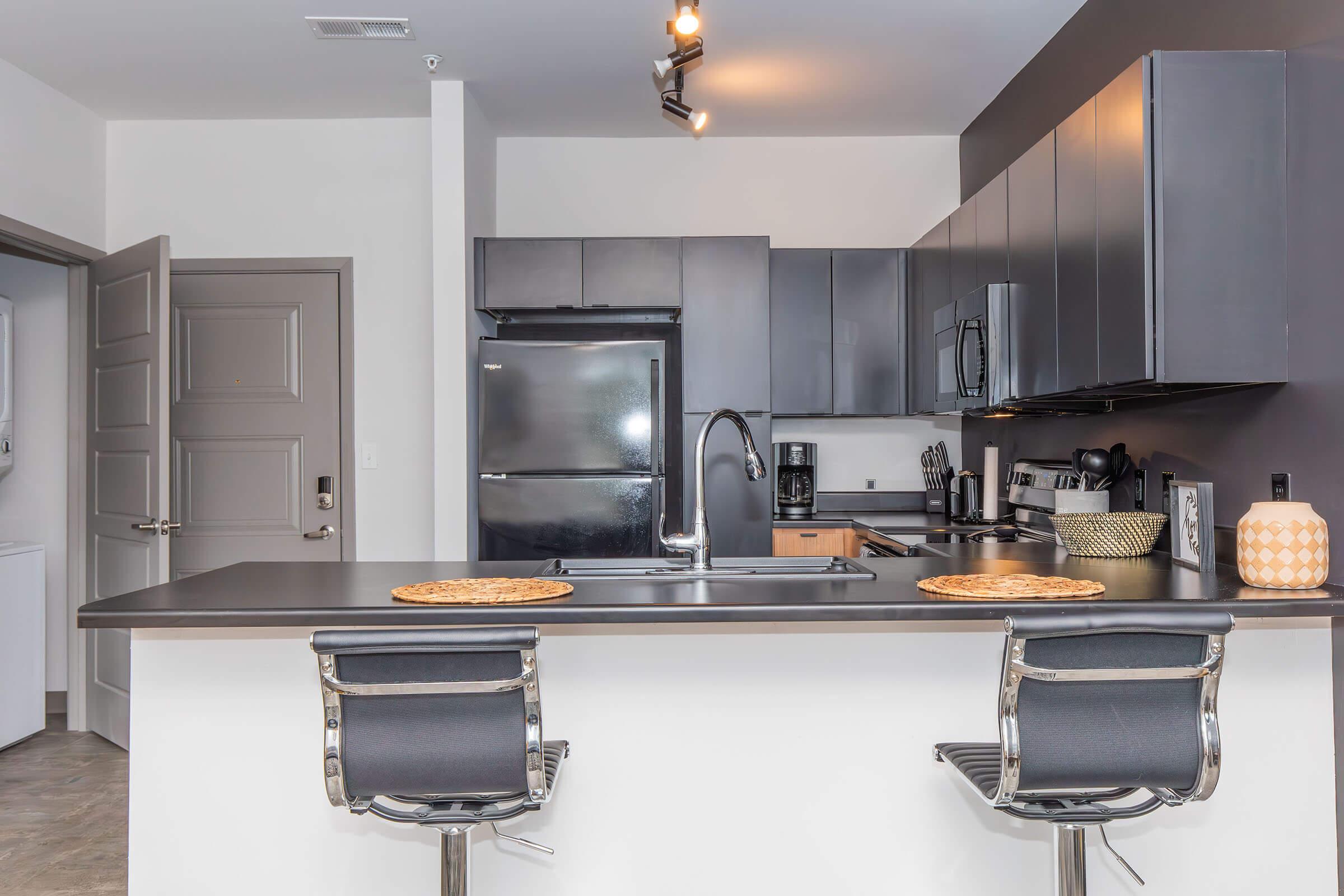 A modern kitchen featuring dark cabinetry, stainless steel appliances, and a black refrigerator. The kitchen island includes two sleek bar stools and decorative plates. Soft lighting enhances the contemporary atmosphere, with an open layout leading to an adjacent door.