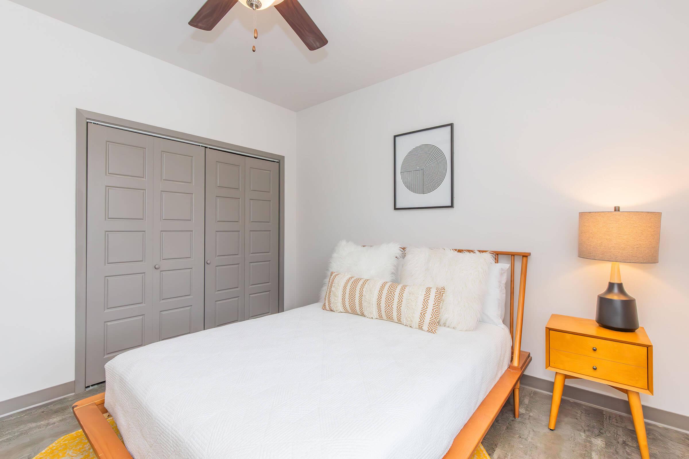 A neatly decorated bedroom featuring a wooden bed with white bedding and fluffy pillows, a wooden nightstand with a lamp, and a framed artwork on the wall. The room has gray closet doors and a soft area rug, creating a cozy and inviting atmosphere.