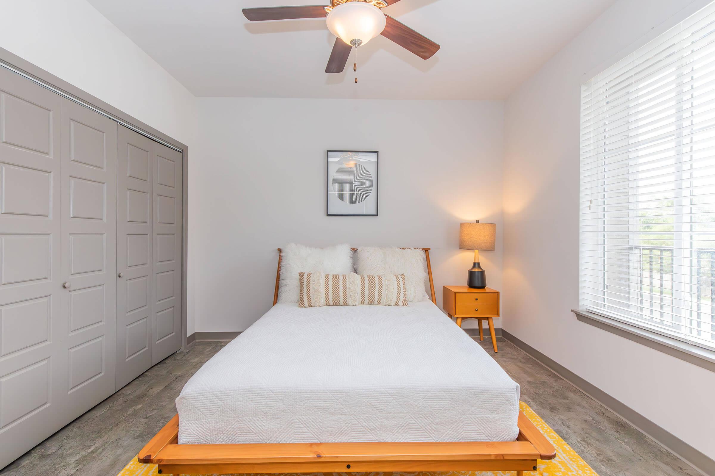 A modern bedroom featuring a wooden bed with a white mattress, two decorative pillows, and a nightstand with a lamp. The walls are light-colored, and there are two closet doors. A window with blinds allows natural light to enter the room, enhancing the bright and airy atmosphere.