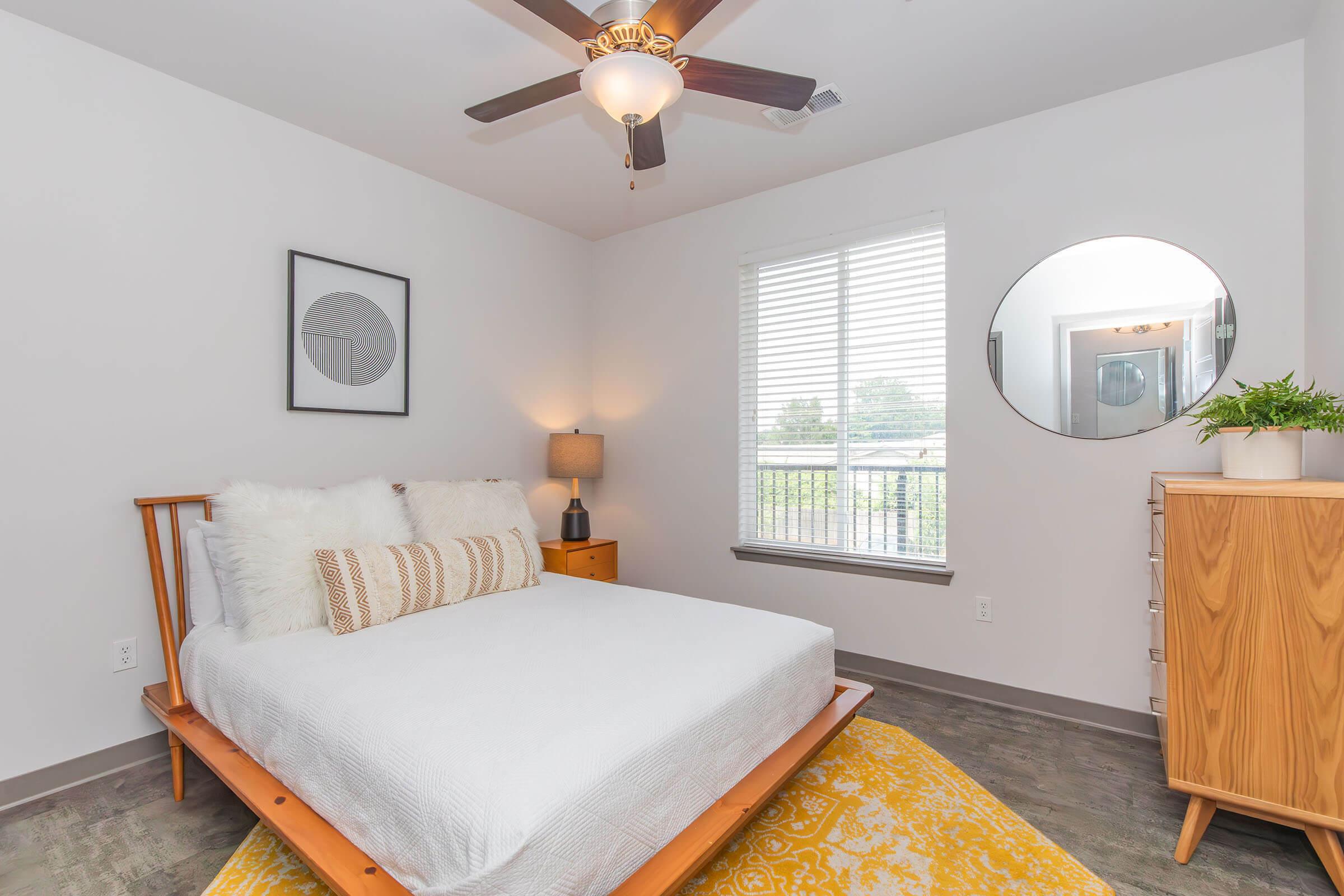 A cozy bedroom featuring a wooden bed with white bedding, a bedside lamp, a round mirror on the wall, and a dresser. Natural light streams in through a window with blinds, illuminating a yellow area rug on the floor. Minimalist decor creates a fresh, inviting atmosphere.