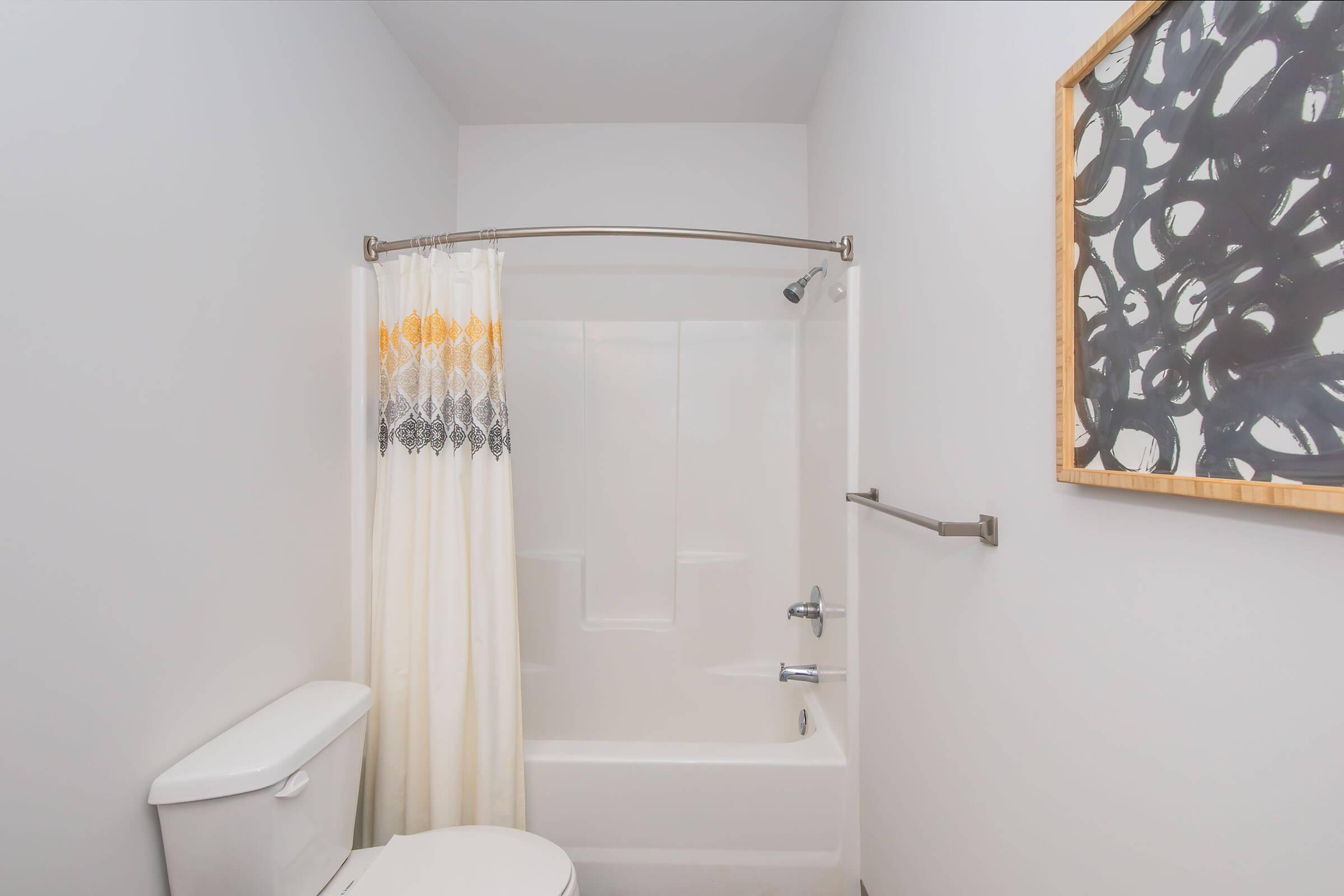A clean, well-lit bathroom featuring a bathtub with a shower, a white curtain with decorative trim, a toilet, and a minimalist wall art piece in a wooden frame. The walls are painted a light color, creating a spacious ambiance.