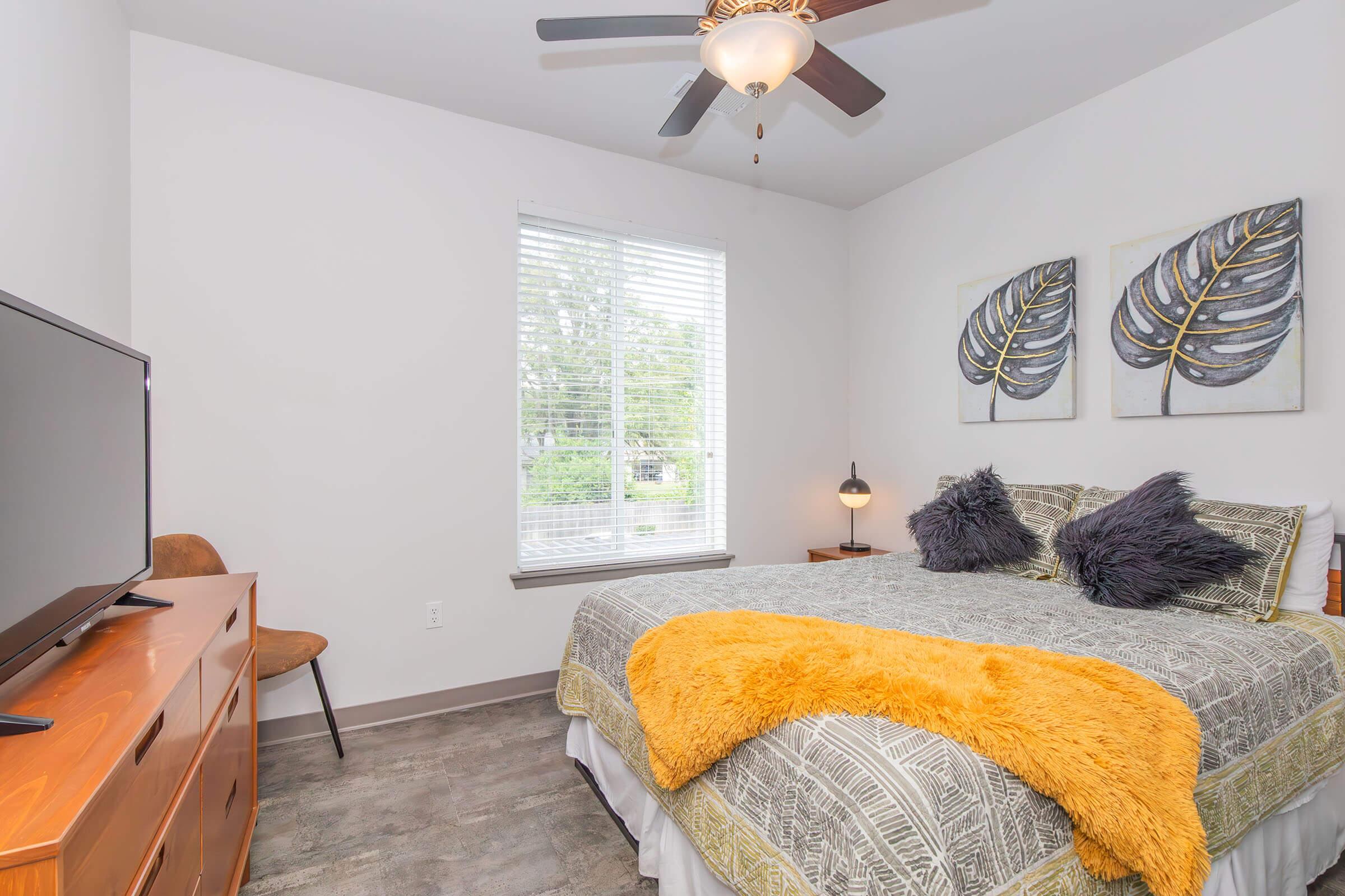 Bright and cozy bedroom featuring a queen-sized bed with a decorative yellow throw blanket. The room includes a wooden dresser, a flat-screen TV, and a chair. Two framed leaf prints adorn the walls, and natural light streams through a large window with white blinds.
