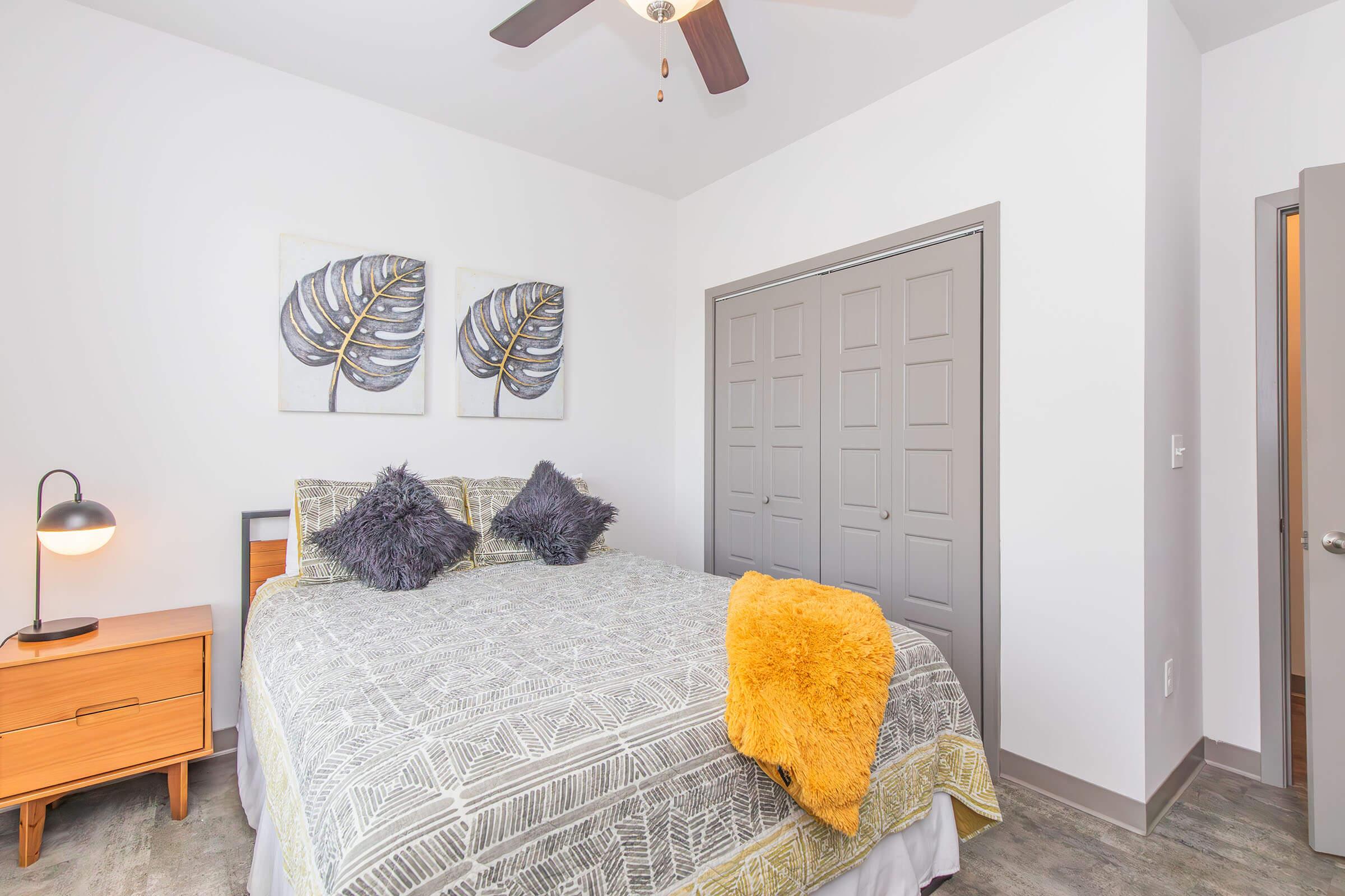 A cozy bedroom featuring a queen-sized bed with a patterned gray and yellow blanket, decorative pillows, and two framed leaf prints on the wall. There’s a stylish nightstand with a lamp and a closet with gray double doors. Natural light highlights the modern design and warm ambiance.
