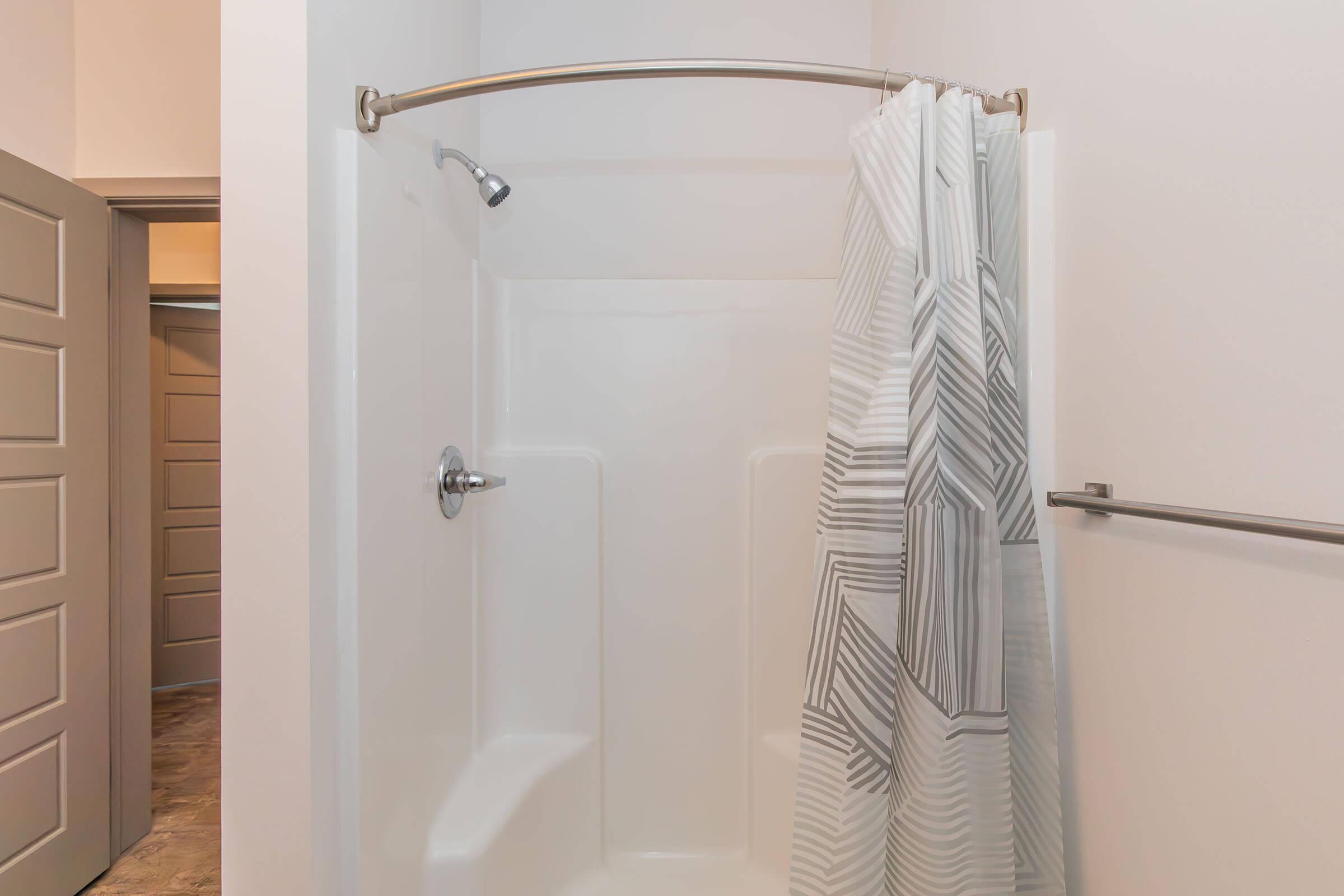 A clean and modern shower area featuring a white bathtub and a curved metal shower rod. A patterned shower curtain partially covers the entrance, and a chrome showerhead is visible. The background shows a closed door with a textured panel. The overall space is well-lit and minimalist.