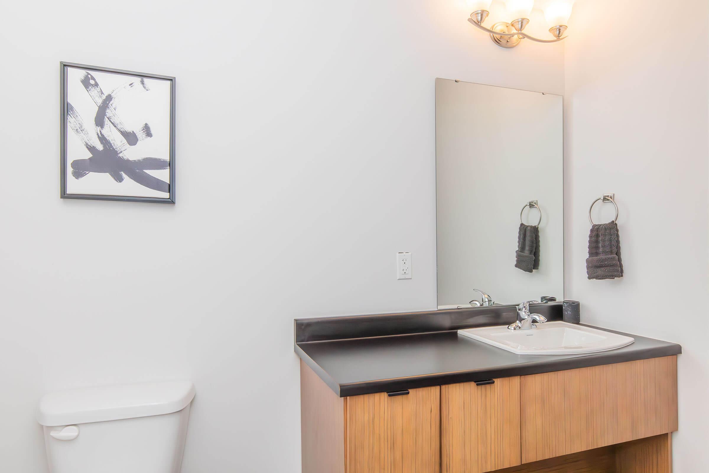 A modern bathroom featuring a wall-mounted mirror above a minimalist sink with a dark countertop. To the left, there is a white toilet, and on the wall, a black and white abstract painting. Two dark towels hang from a towel rack beside the sink. The overall decor is clean and contemporary.