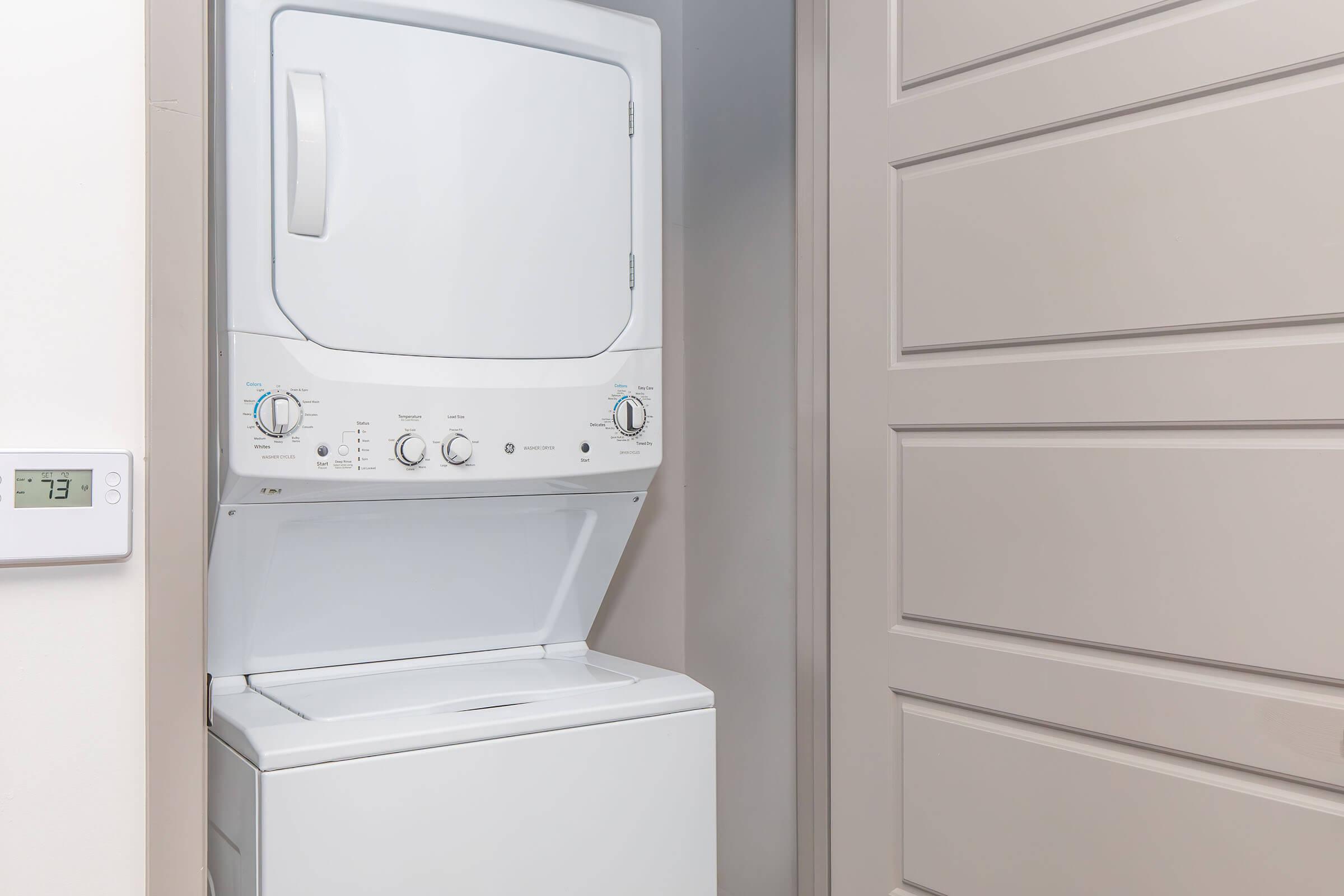 A stacked washer and dryer unit in a minimalist laundry space, with a light-colored wall in the background. A thermostat displays a temperature reading of 73 degrees on the left side. The door beside the unit is a simple design with horizontal paneling.