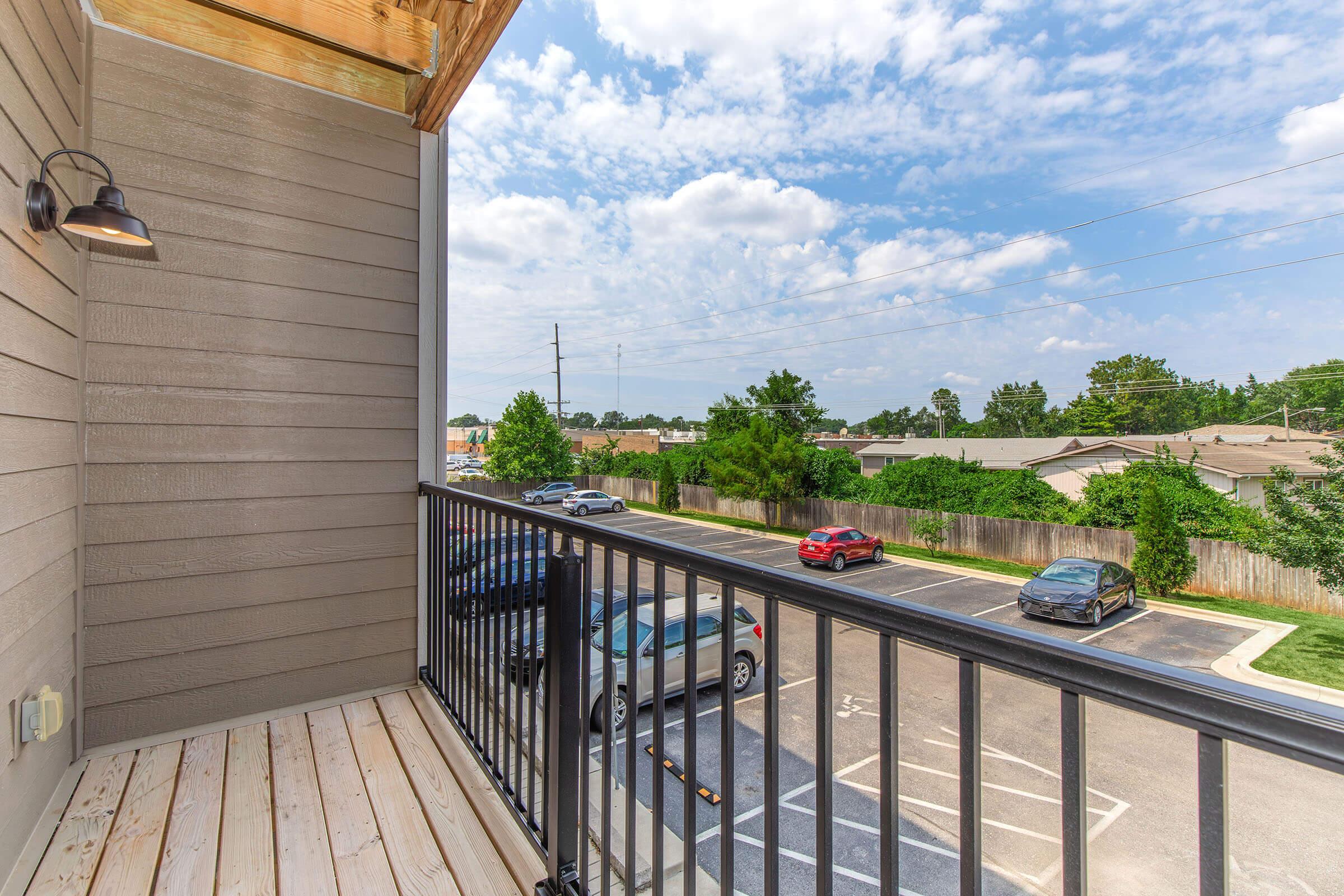 View from a balcony overlooking a parking lot and residential area. The scene features blue skies with scattered clouds, green trees, and parked cars. The balcony has wooden flooring and a metal railing, with a light fixture mounted on the wall.