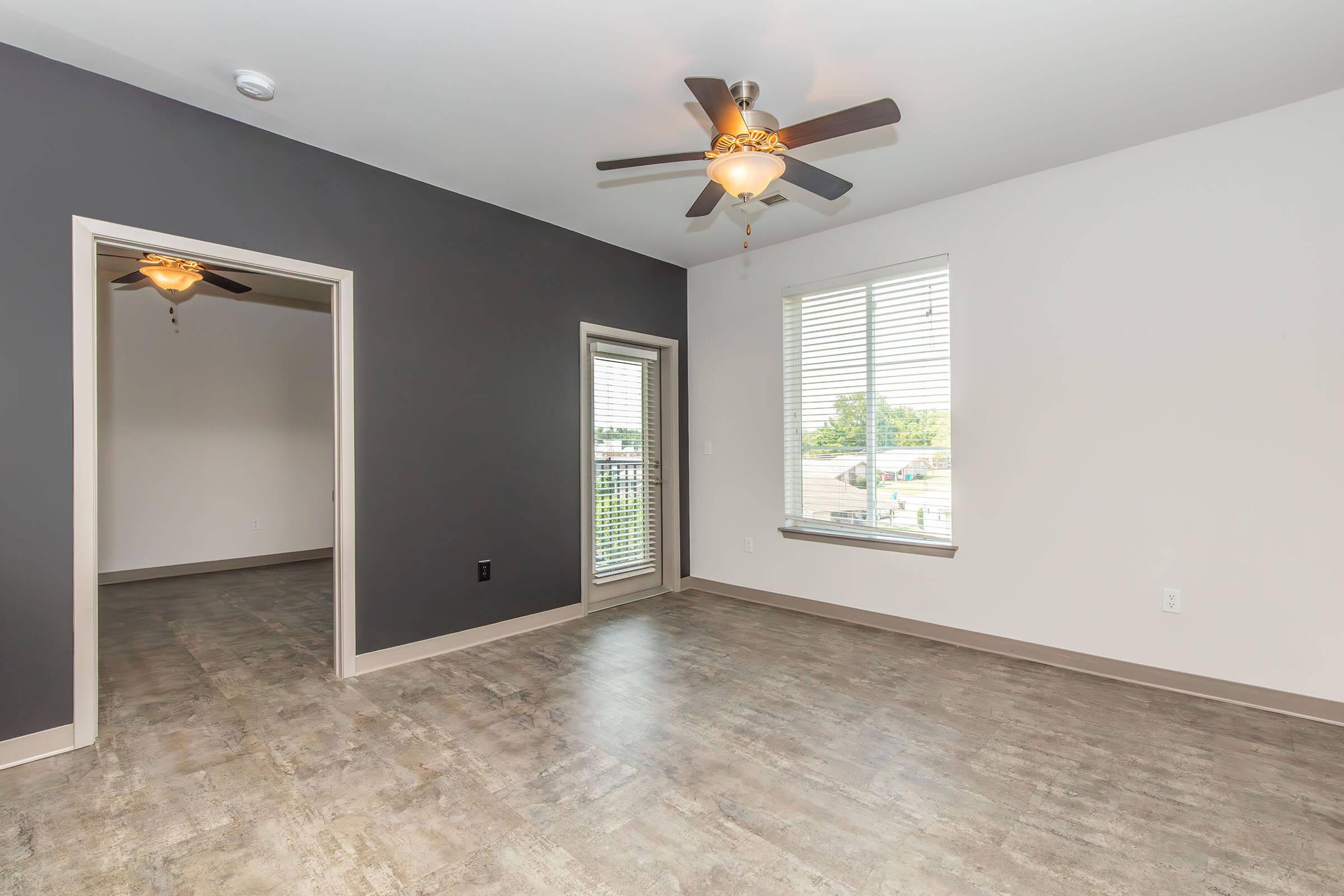 A spacious room featuring a gray accent wall and light hardwood flooring. The ceiling has a ceiling fan with light fixtures. A window with blinds illuminates the space, and there is a door leading to an outdoor area. Another room is visible through the open doorway across the room.