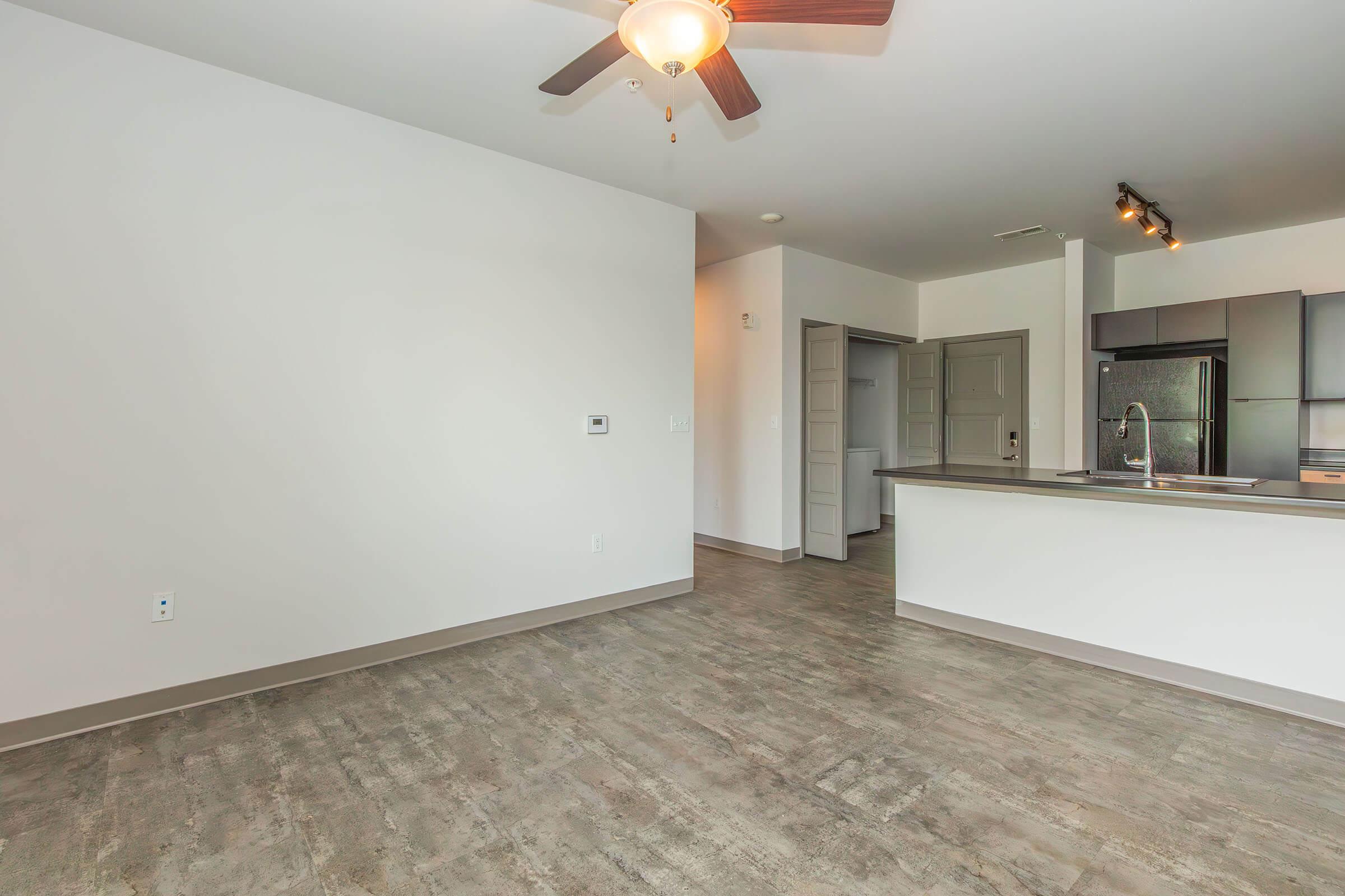 A modern, empty apartment interior featuring a ceiling fan, light wood-style flooring, and open layout. The kitchen with dark cabinets is visible in the background, along with a doorway leading to another room. Walls are painted light, creating a spacious feel.
