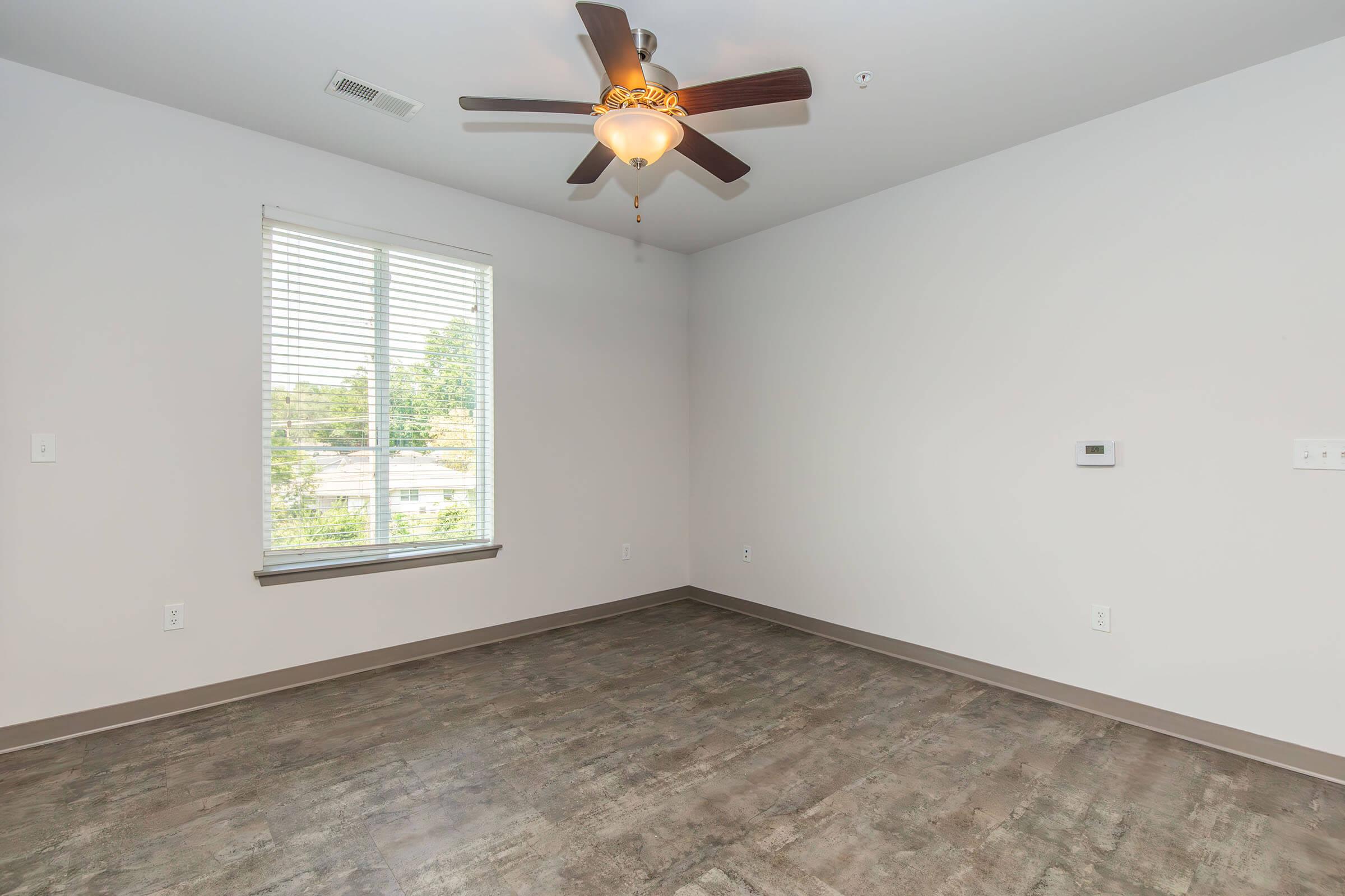 Empty room with light gray walls and a ceiling fan. A window with white blinds allows natural light in, and the floor features a modern, wood-look design. An air conditioning unit is mounted on the wall, contributing to the spacious and minimalistic feel of the space.