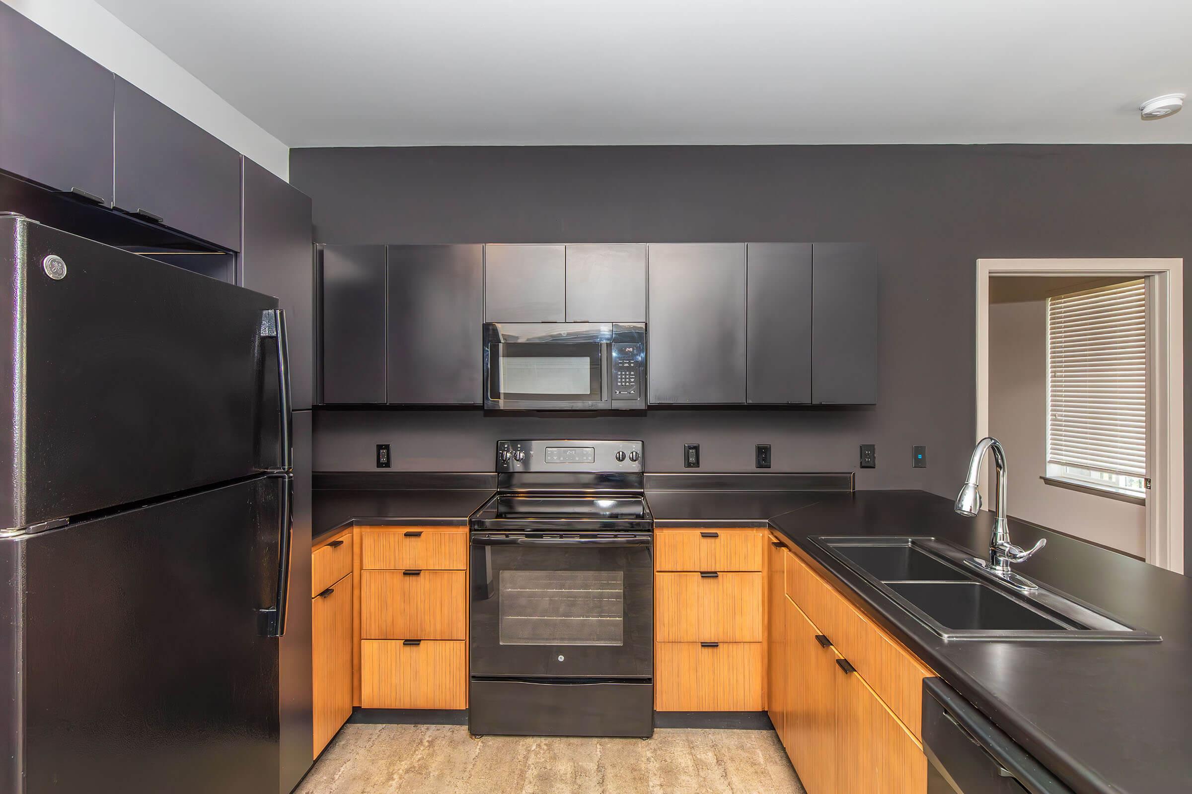 Modern kitchen featuring black and wood cabinetry, a black refrigerator, a stainless steel microwave above a black stove, and a dark countertop. The design is sleek and contemporary, with minimalistic aesthetics and ample storage. Natural light enters from a nearby window, creating a bright atmosphere.
