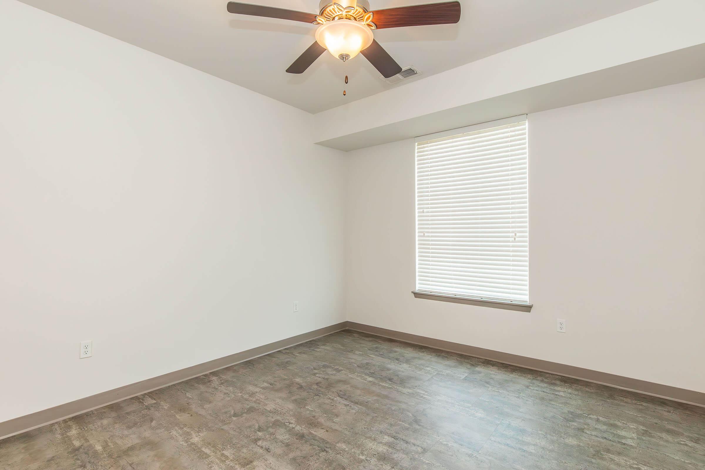Empty room with white walls and a light-colored wood floor. A ceiling fan with lights is centered above the room, and there is a window on the left with blinds. The overall space is bright and minimalistic, creating an open and airy atmosphere.