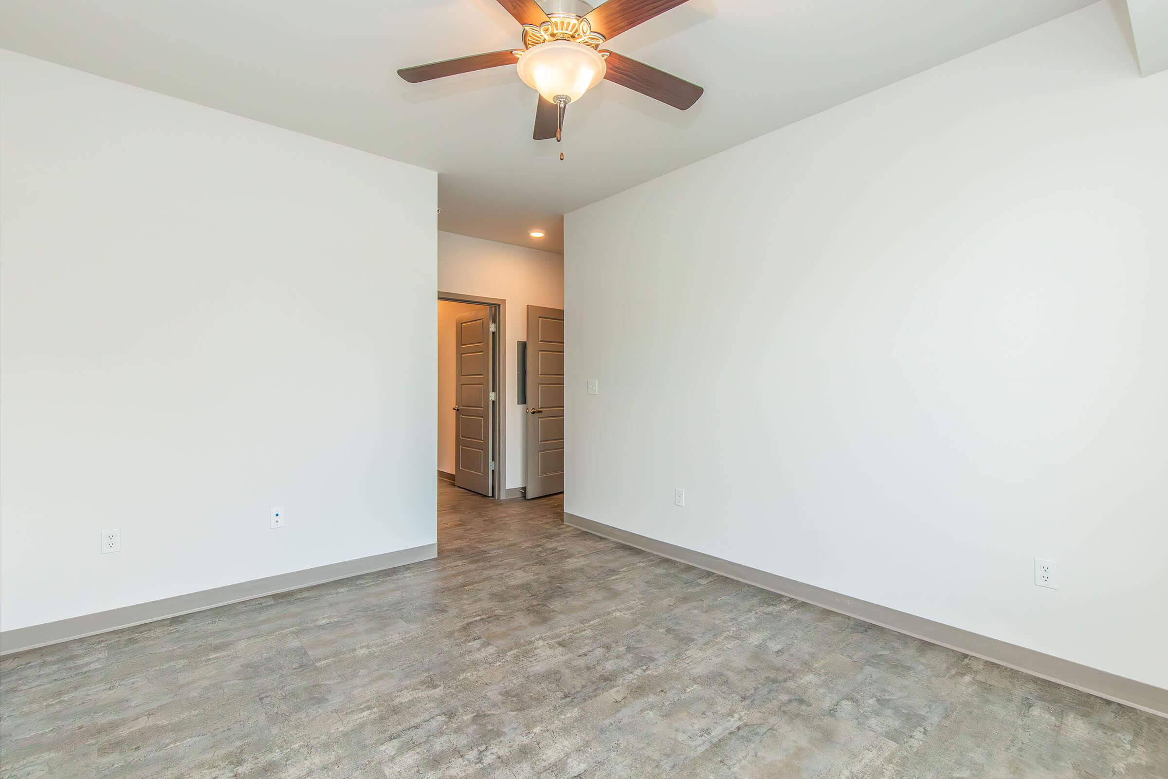 Empty room with light-colored walls and a ceiling fan. The floor has a modern finish, and there's an open doorway leading to another room. Natural light enters from the doorway, creating a bright and inviting space.