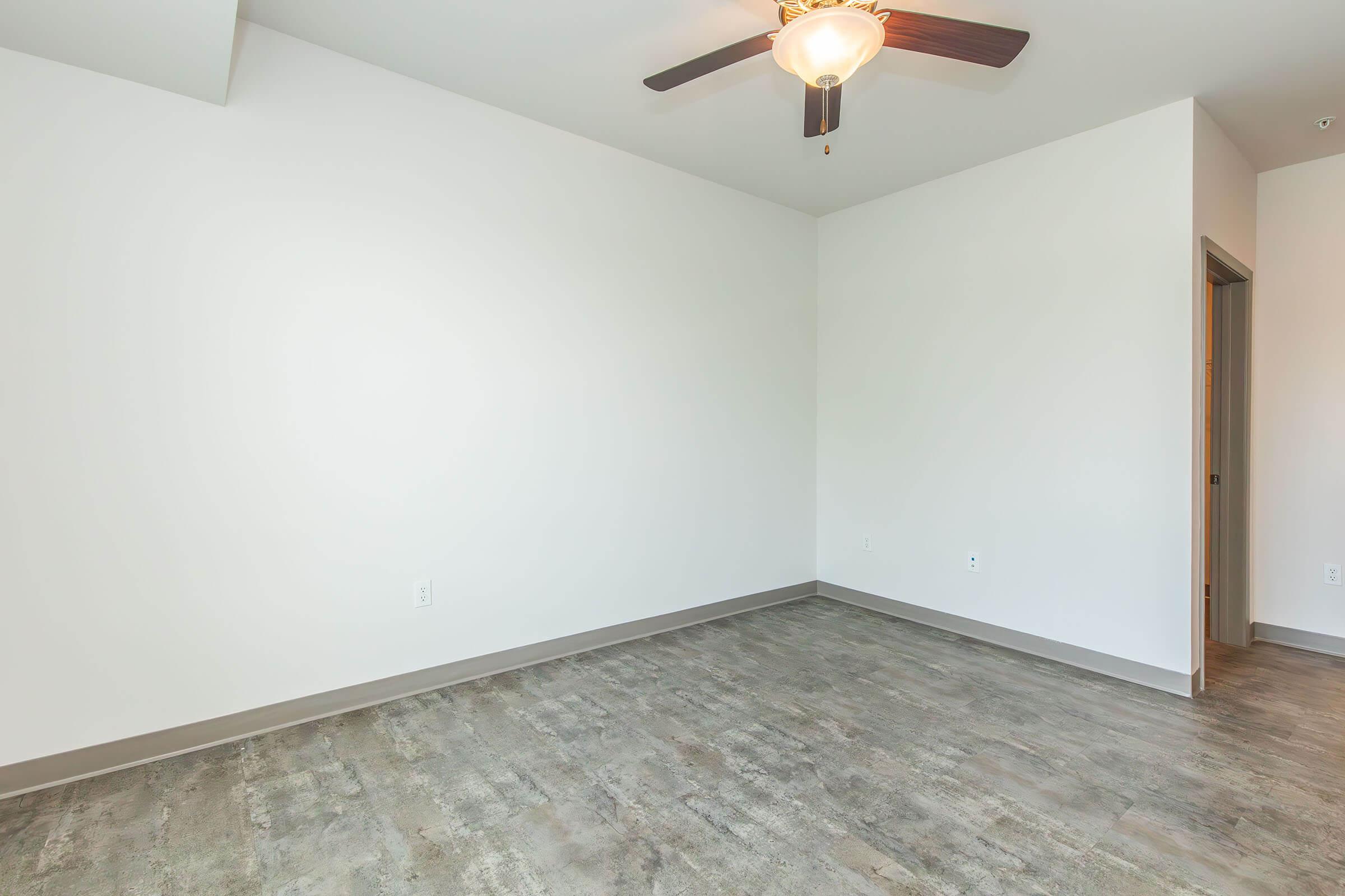 Empty room with light-colored walls and a ceiling fan. The floor has a wood-like finish, and there is a doorway on the right leading to another space. Natural light is coming from a window, creating a bright and open atmosphere.