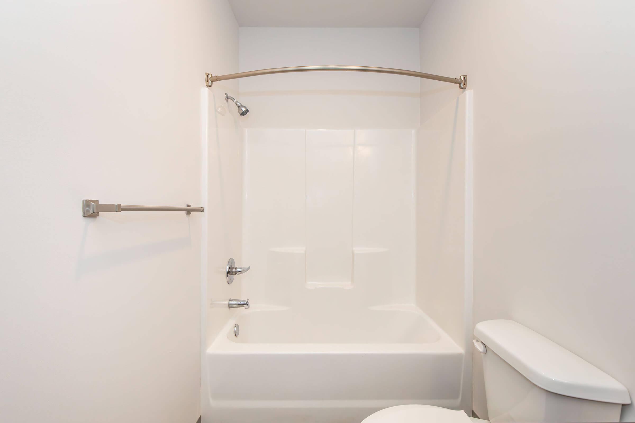 A clean, minimalist bathroom featuring a white bathtub with a shower, a chrome showerhead, and a towel bar. The walls are plain and light-colored, giving a spacious look. A white toilet is positioned beside the bathtub, contributing to the simple design of the space.
