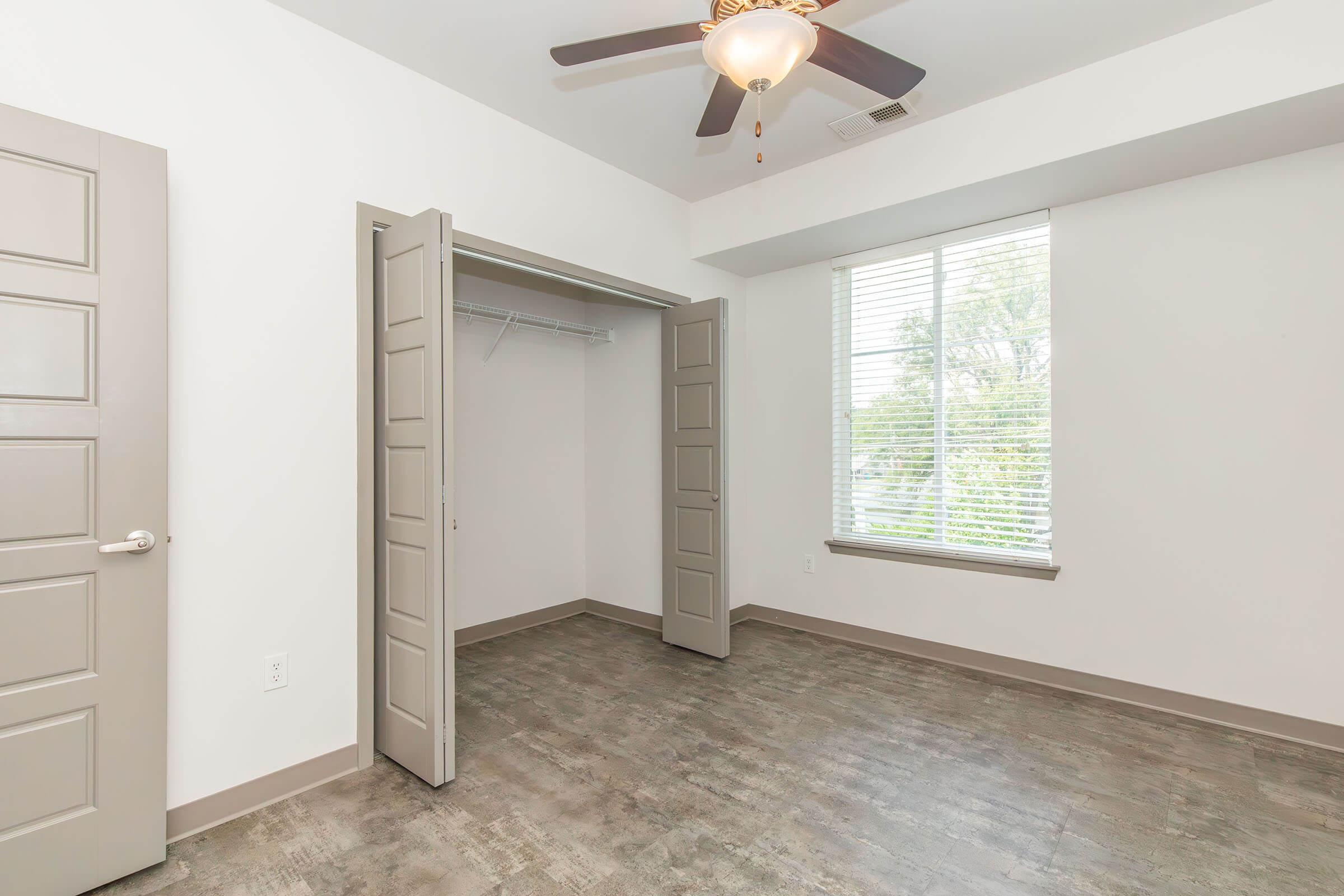 Empty room featuring light-colored walls, a ceiling fan, and a large window with blinds. The closet has double doors and is positioned on one side of the room. The floor has a modern, textured design, contributing to a spacious and bright atmosphere.