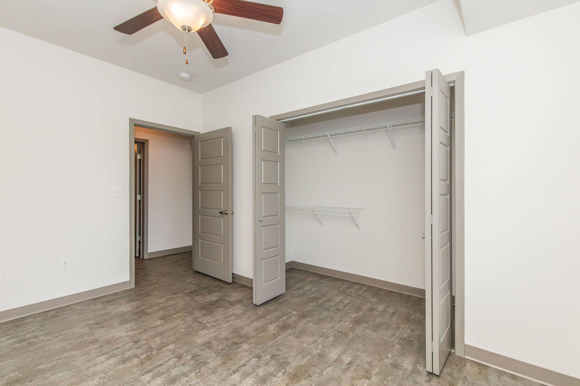 An empty room featuring a ceiling fan, light fixture, and light-colored walls. Two open closet doors reveal a spacious closet with wall-mounted shelving. The floor is wooden and has a neutral tone, enhancing the room's bright and airy feel.