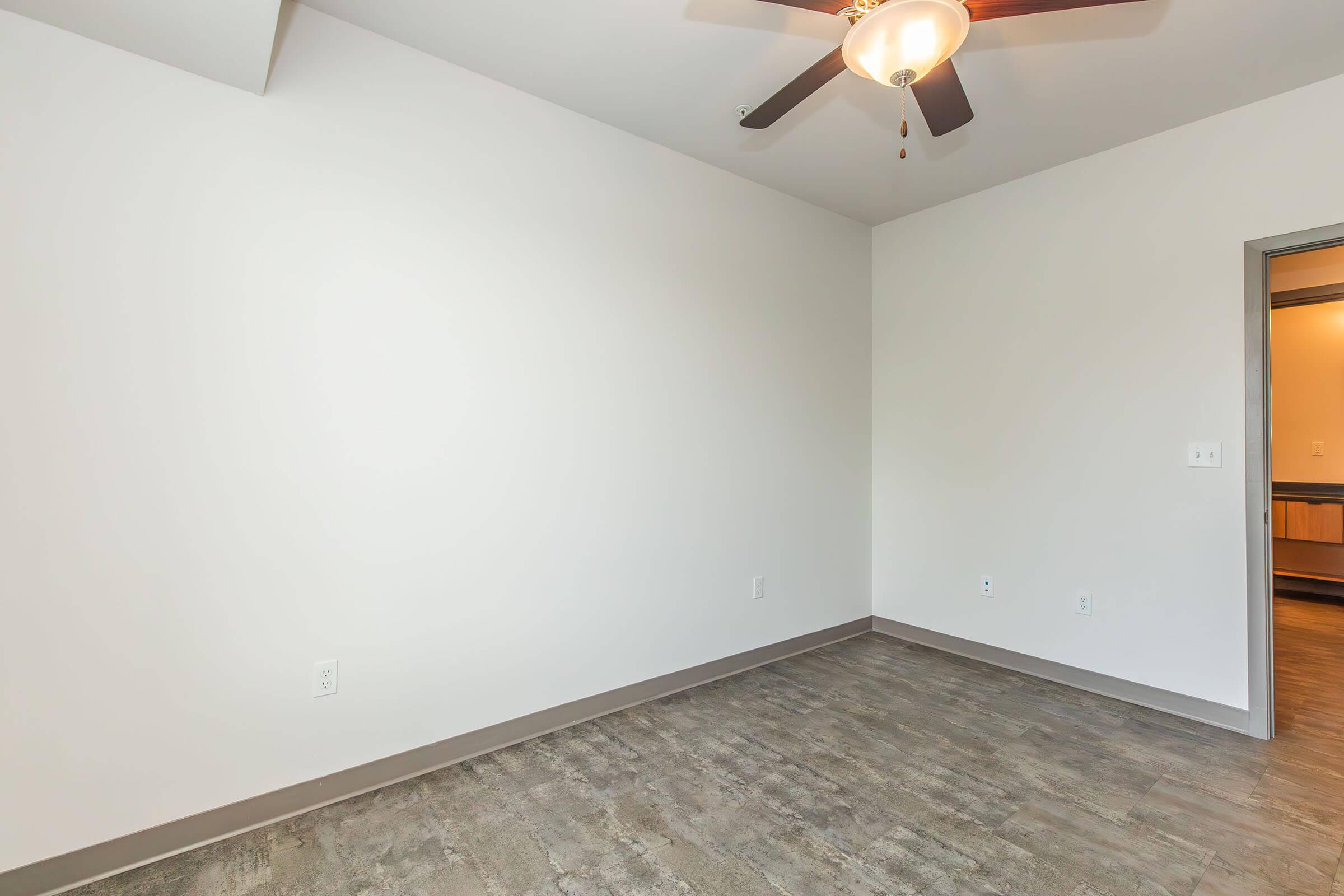 A clean, empty room with light-colored walls, a ceiling fan, and a gray floor. A doorway leading to another space is visible on the right, while the rest of the room features no furniture or decorations, creating a spacious and minimalist feel. Natural light brightens the space.