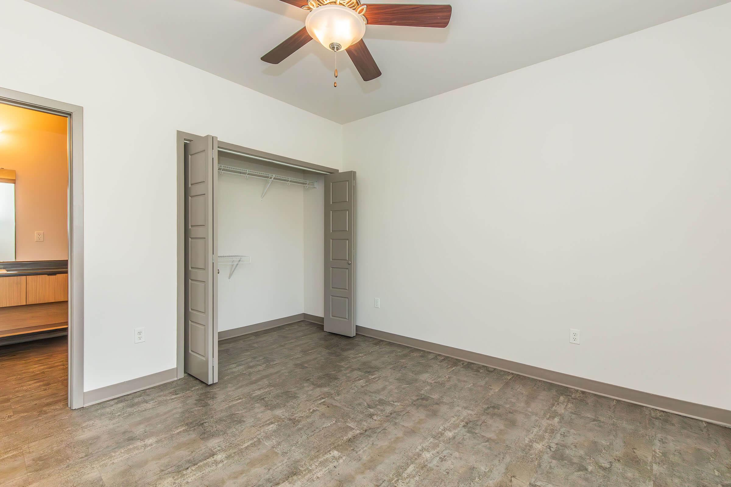 A bright, empty room featuring a ceiling fan and light fixture. Light-colored walls and a wooden floor create a spacious feel. There's an open closet with sliding doors on one side and a doorway leading to another area, suggesting a well-lit, modern living space.
