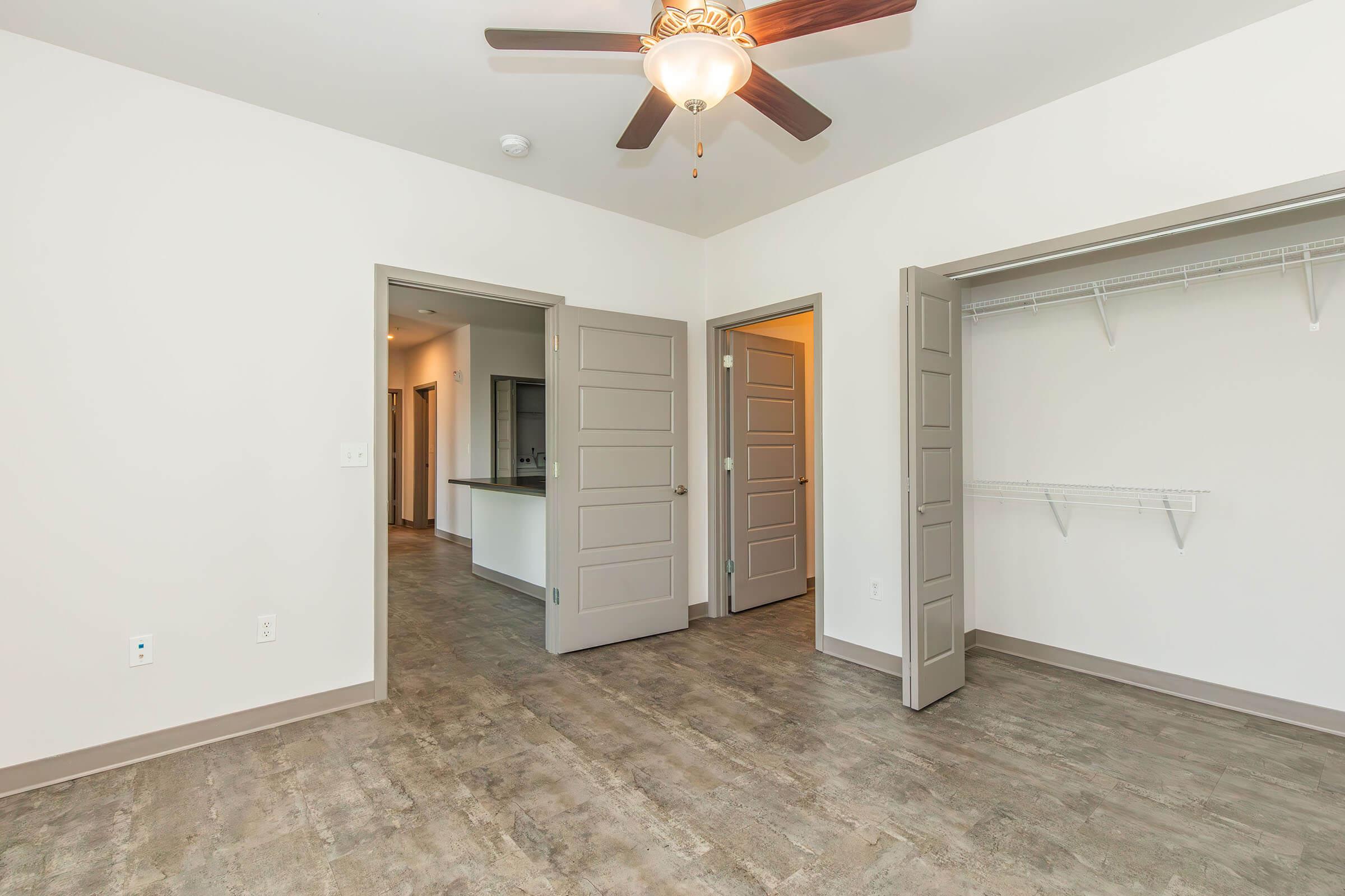 Interior view of a modern bedroom featuring a ceiling fan, light-colored walls, and laminate flooring. Two doors lead to different areas, one open to an adjacent room and the other to a closet with shelving. The layout appears spacious and well-lit, with a neutral color palette.