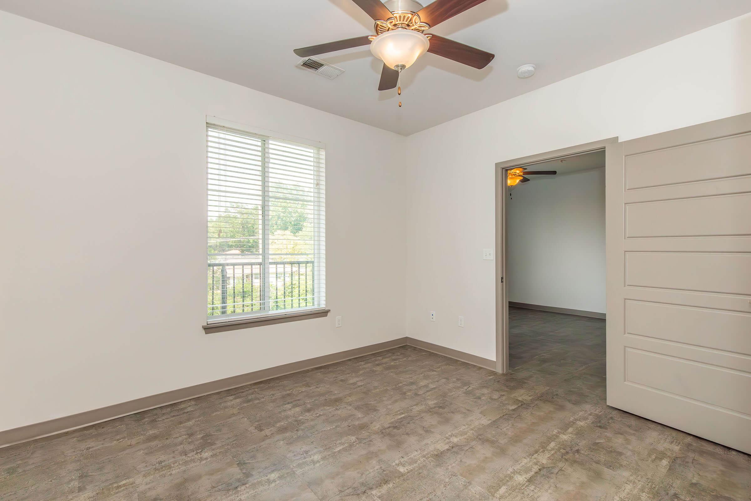 A well-lit interior of an empty room featuring a ceiling fan, a window with blinds, light-colored walls, and a door leading to another space. The floor has a modern, textured finish, creating a spacious and inviting atmosphere.