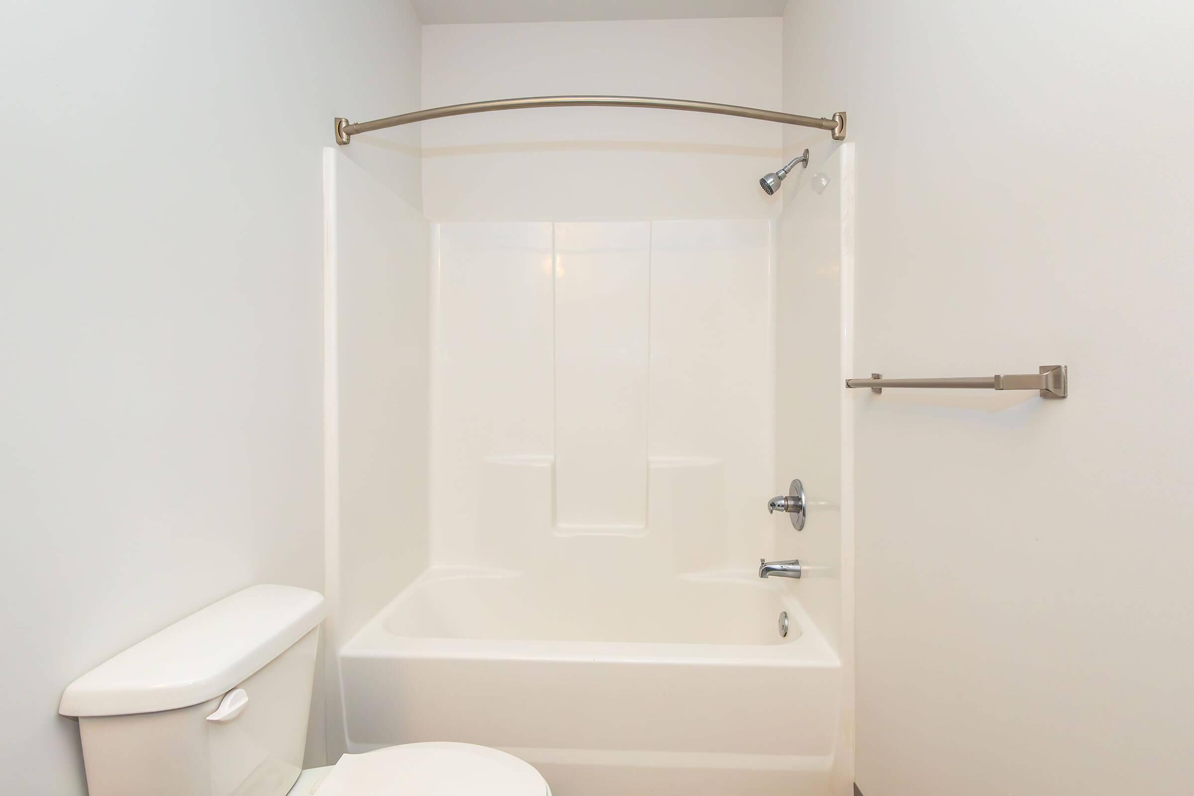 A clean, modern bathroom with a white bathtub and shower combination, white toilet, and a towel bar. The walls are plain and light-colored, creating a simple and spacious appearance.