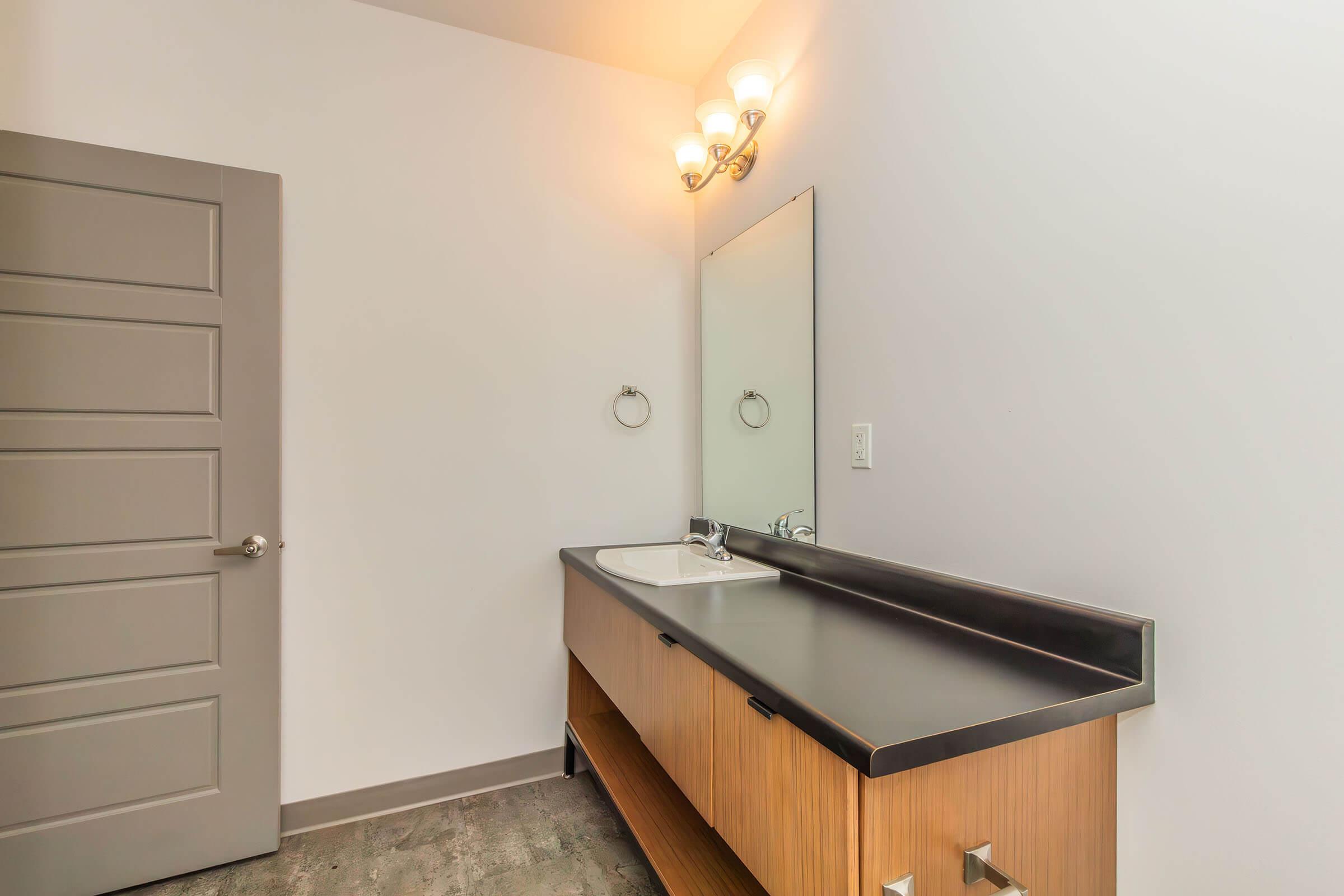 A modern bathroom featuring a sleek wooden vanity with a sink, a large mirror above it, and a light fixture with three bulbs. The walls are painted white, and a gray door is visible on the left side of the image. The flooring appears to be a light-colored tile.