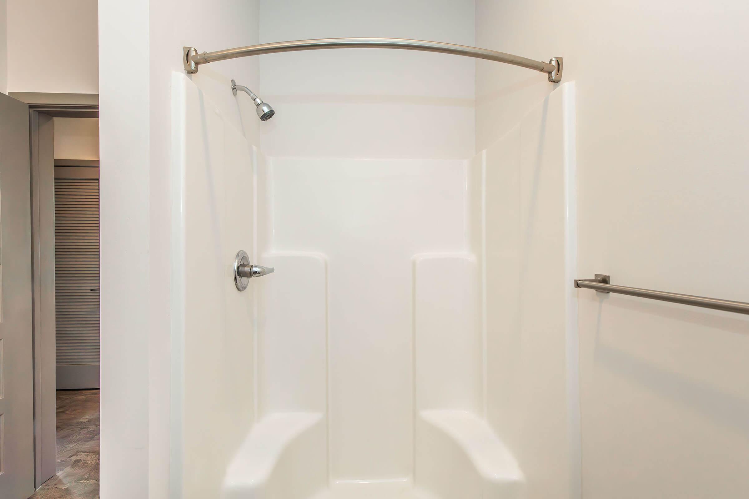 A clean, modern shower stall with a white fiberglass interior, featuring a handheld showerhead mounted on a curved chrome rod. The walls are smooth and minimalistic, enhancing the bright, open feel of the space. A grab bar is installed for safety, and there's an adjoining wall visible in the background.