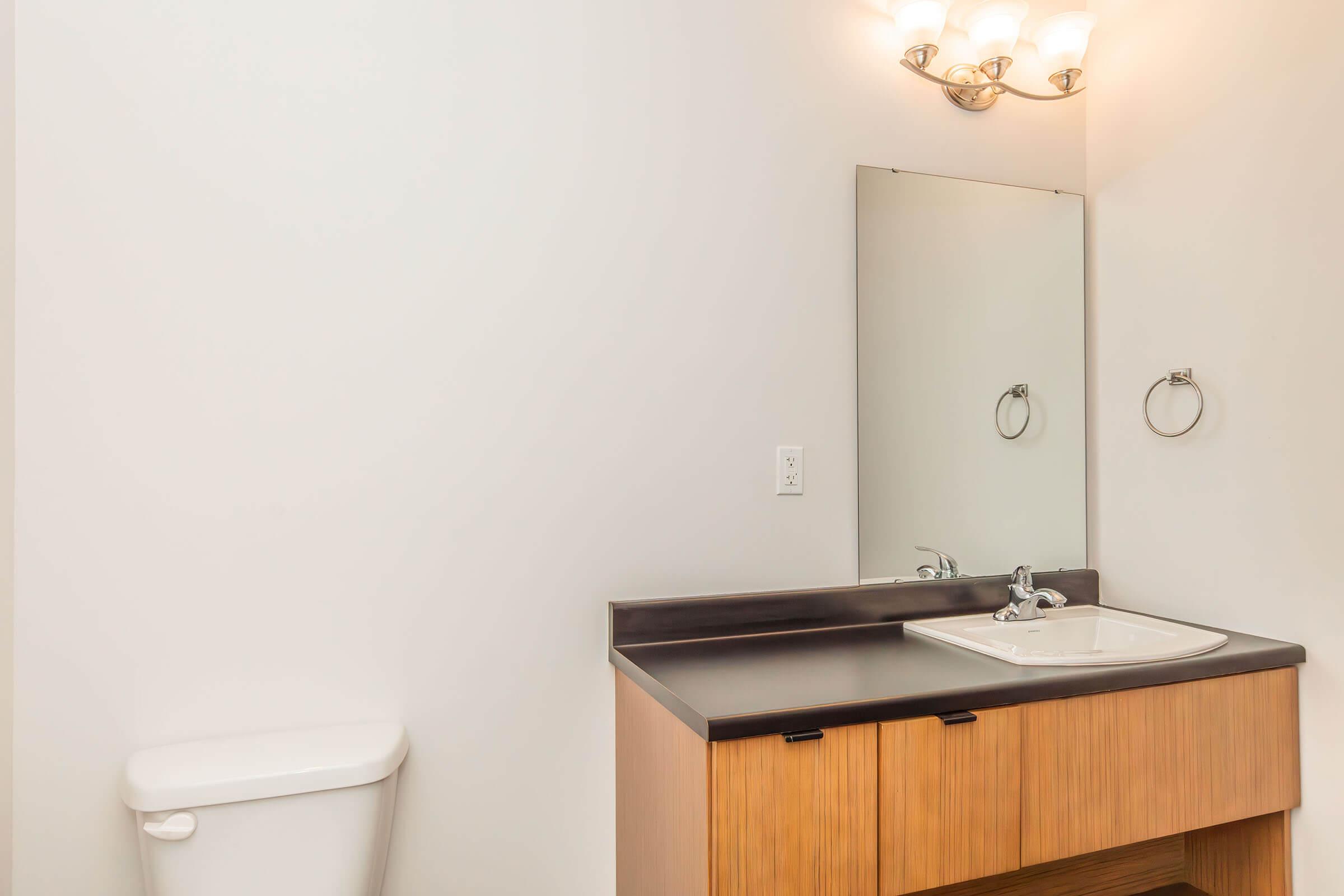 A clean and modern bathroom featuring a white toilet, a wooden vanity with a black countertop, a sink, and a wall-mounted mirror. There is a towel ring next to the sink, and the walls are painted in a neutral color, providing a bright and spacious feel. Soft lighting is provided by a light fixture above the mirror.