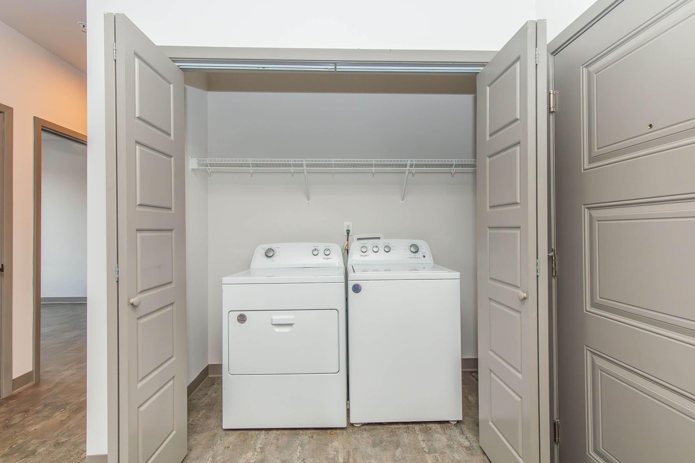 A laundry closet featuring a white washing machine and dryer side by side, with a wire shelf above for storage. The closet doors are wide open, revealing a clean and modern interior with light-colored walls and a gray floor.