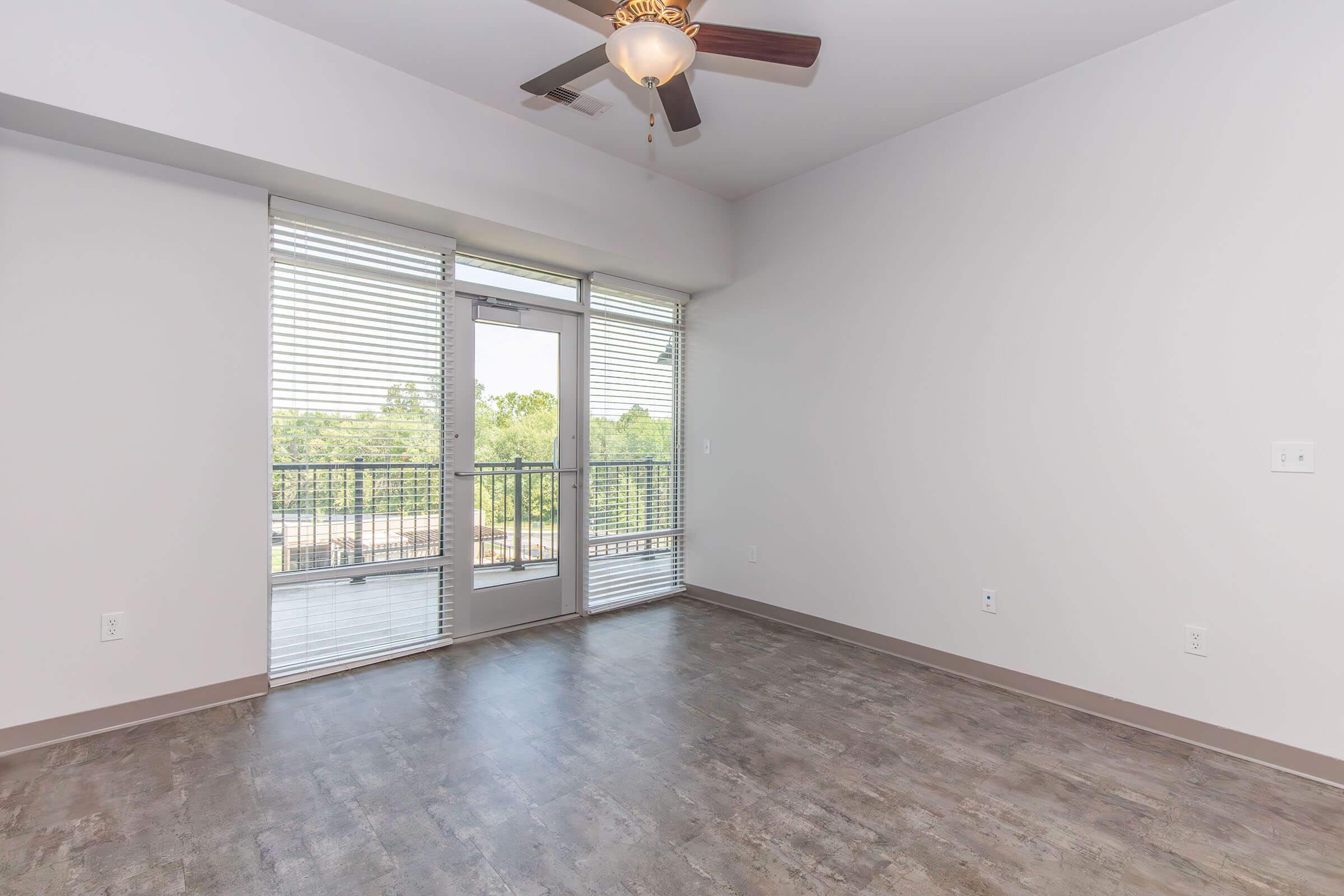 A spacious, empty living room with light gray walls and a ceiling fan. Large windows with horizontal blinds allow natural light to fill the room. A glass door leads to a balcony with a view of trees outside. The flooring is a modern, dark laminate, adding to the contemporary aesthetic.