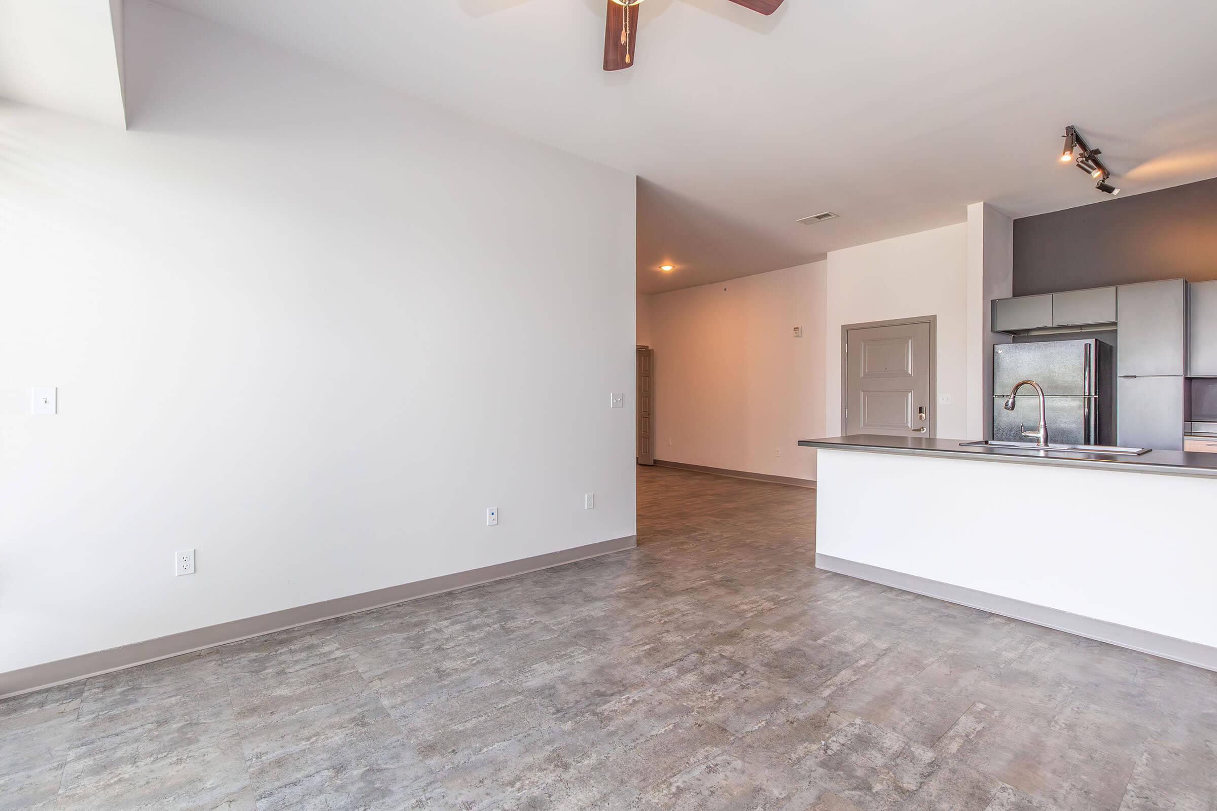Spacious interior of a modern apartment featuring a light gray floor, white walls, and ceiling fan. The kitchen area is open with sleek cabinetry and stainless steel appliances. There is ample natural light coming from the windows, creating a bright and inviting living space.