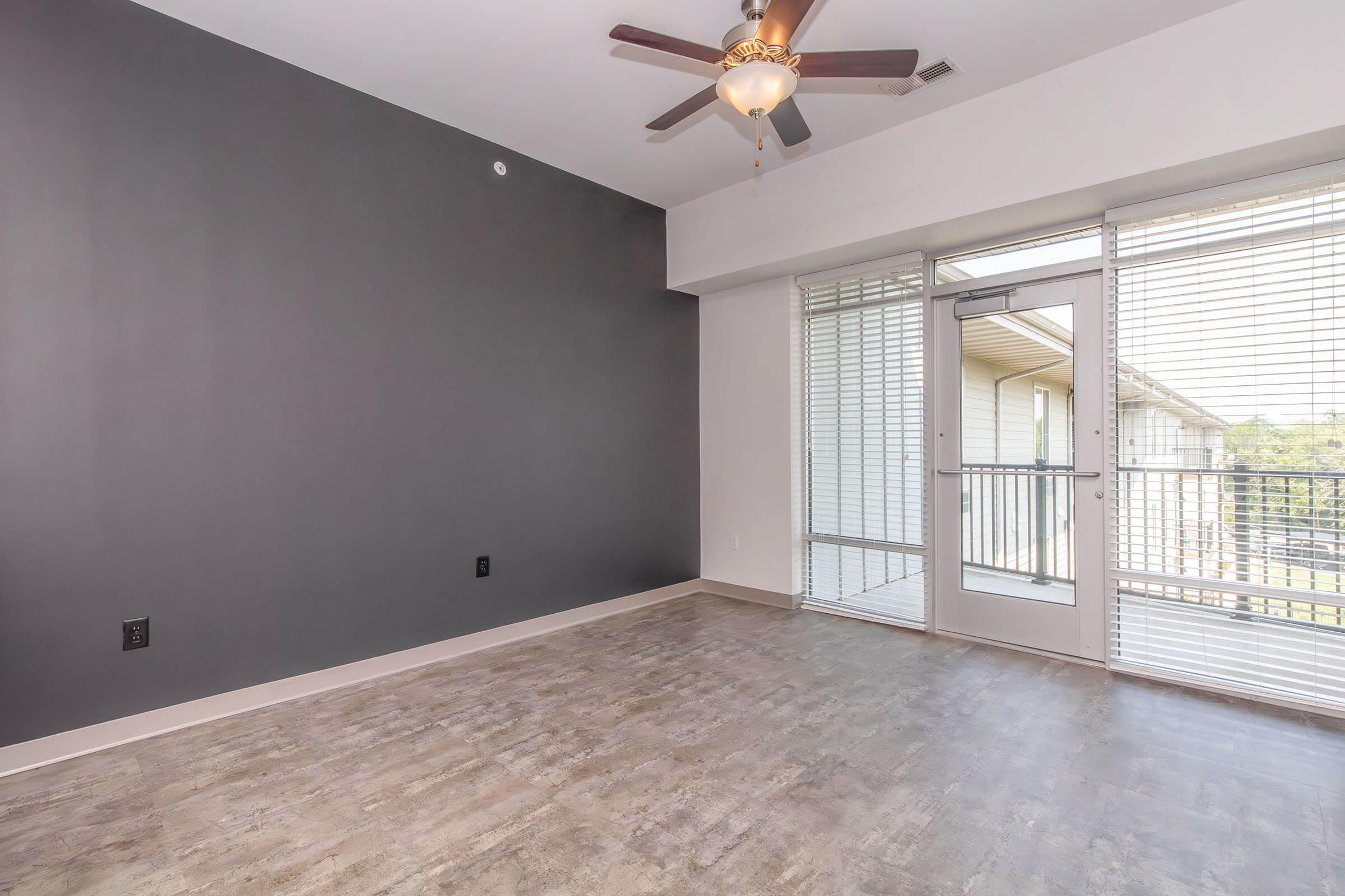 A spacious, modern living area with a gray accent wall, light-colored flooring, and a ceiling fan. Large windows provide natural light and a view of a balcony. The design is minimalistic, creating an open and inviting atmosphere.