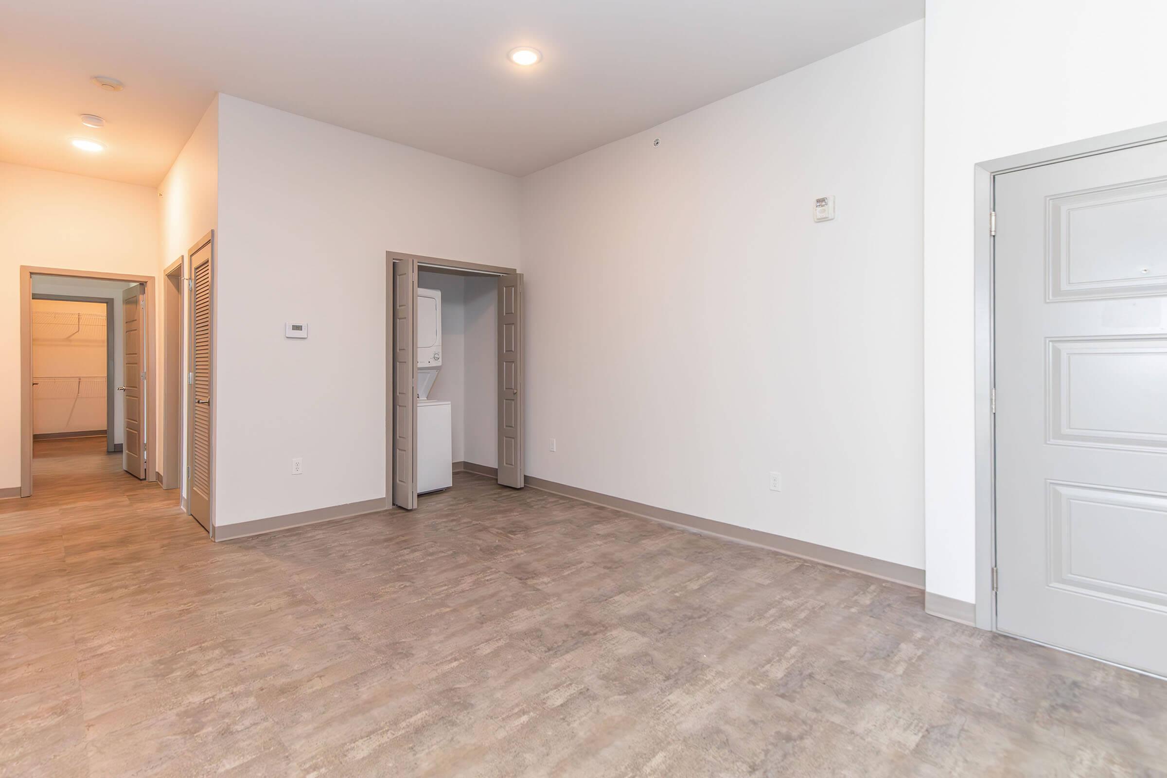 Empty room with light-colored walls and gray flooring. The entrance door is on the right, and there are two open doors leading to a closet and a laundry area. The space has ample natural light, creating a bright and airy atmosphere.