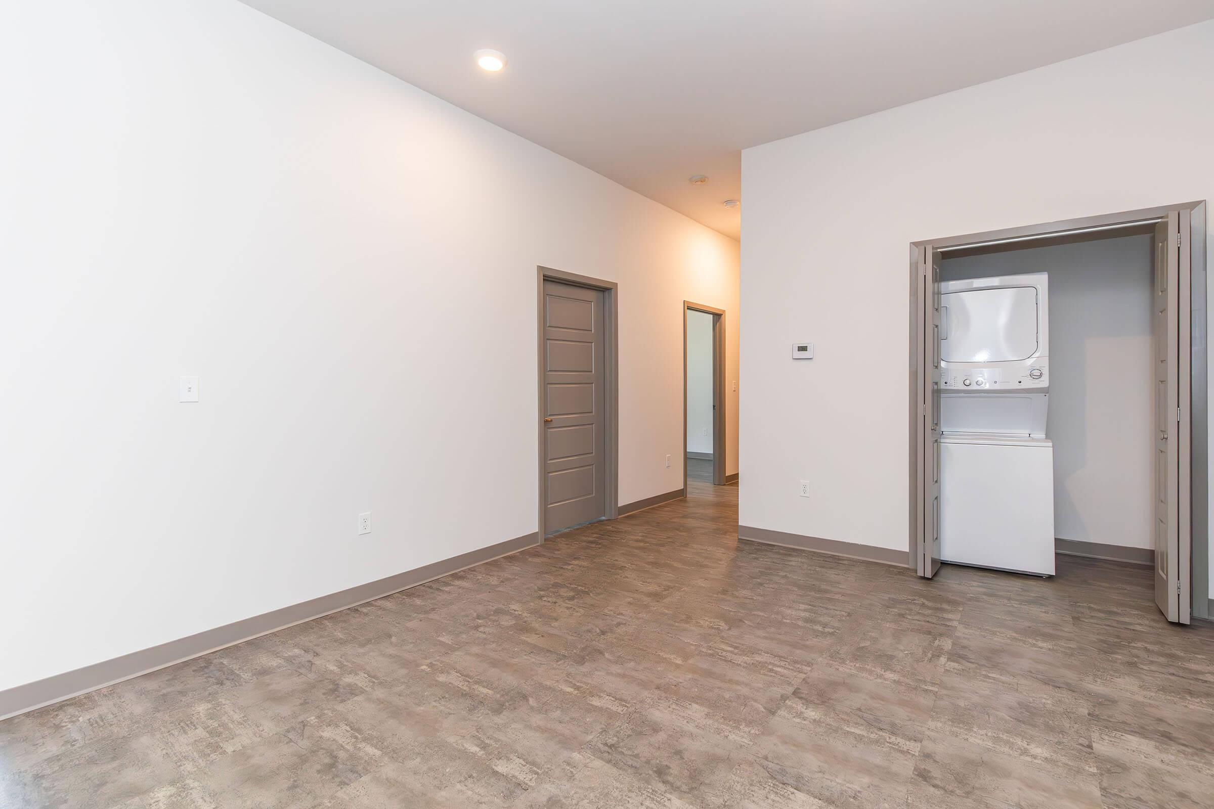 A spacious and modern interior of an apartment, featuring gray walls and flooring. To the left, there is a closed door, while another door leads to a laundry area with a stacked washer and dryer. Natural light is evident, creating a bright and inviting atmosphere.
