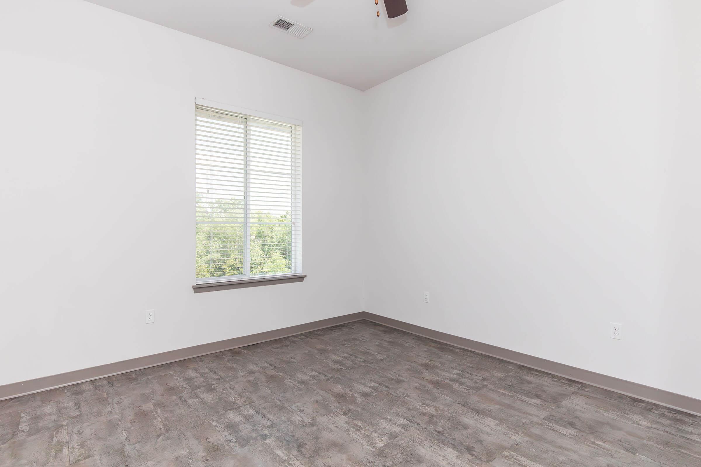 Empty room with light gray walls and a ceiling fan. A window with white blinds allows natural light to enter, revealing a view of greenery outside. The floor features light-colored wood or laminate. The overall atmosphere is spacious and minimalist, suitable for various uses.