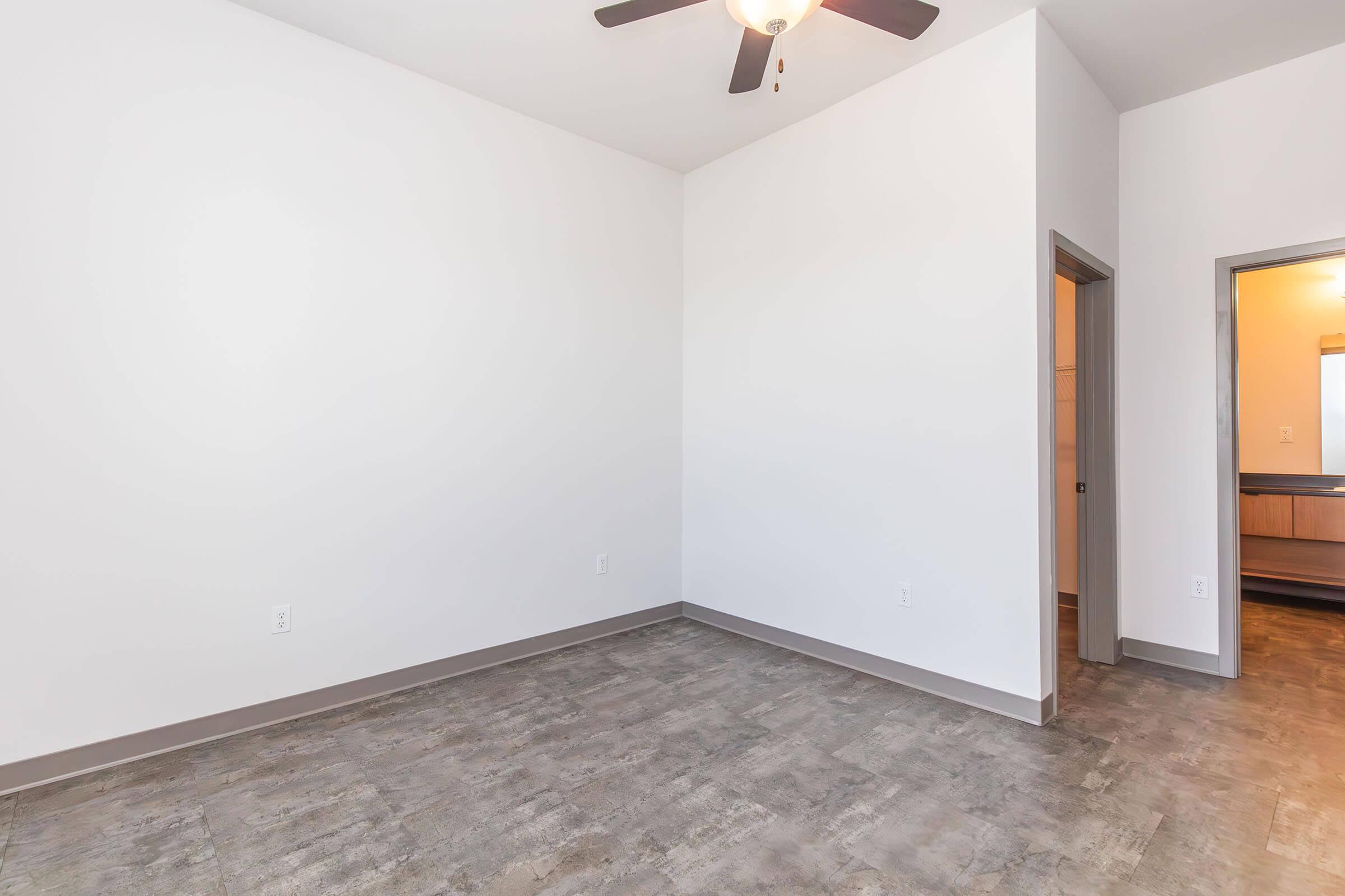 Empty room with light-colored walls, a ceiling fan, and a gray floor. A door leads to another space, and a partially visible area suggests a layout open to the kitchen or bathroom. The room is bright and spacious.
