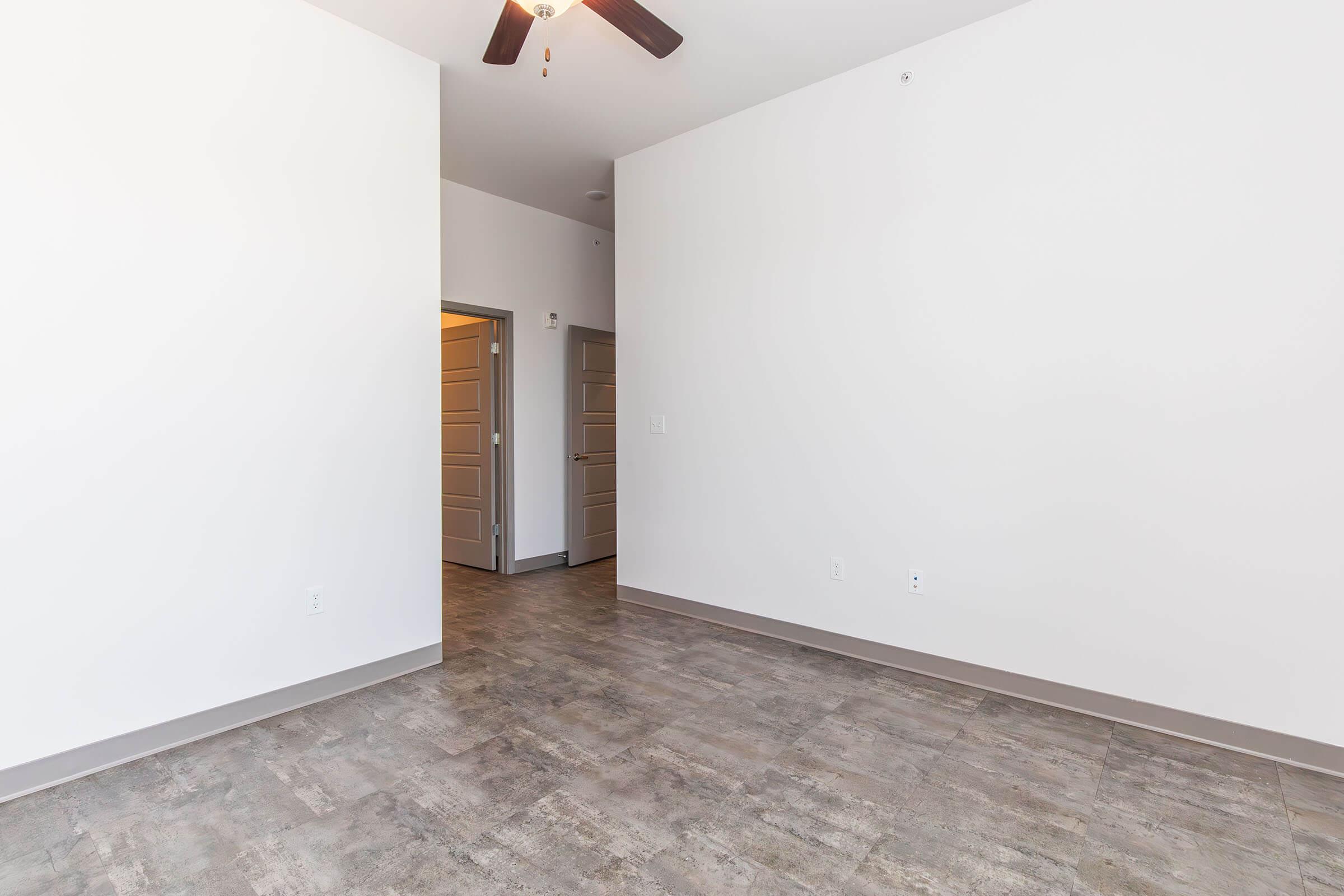 A spacious, empty room with light-colored walls and a modern ceiling fan. The flooring is a sleek, grey tile. A doorway leads to another room, suggesting potential for various uses or decoration. Natural light may brighten the space, creating a welcoming atmosphere.