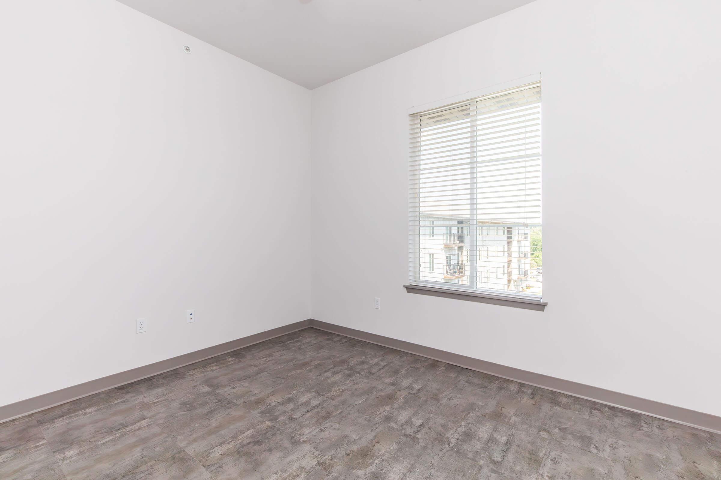 A bare room with light-colored walls and a large window featuring blinds. The floor has a wood-like finish, and there are no furnishings or decorations. The window offers a view of an exterior space.