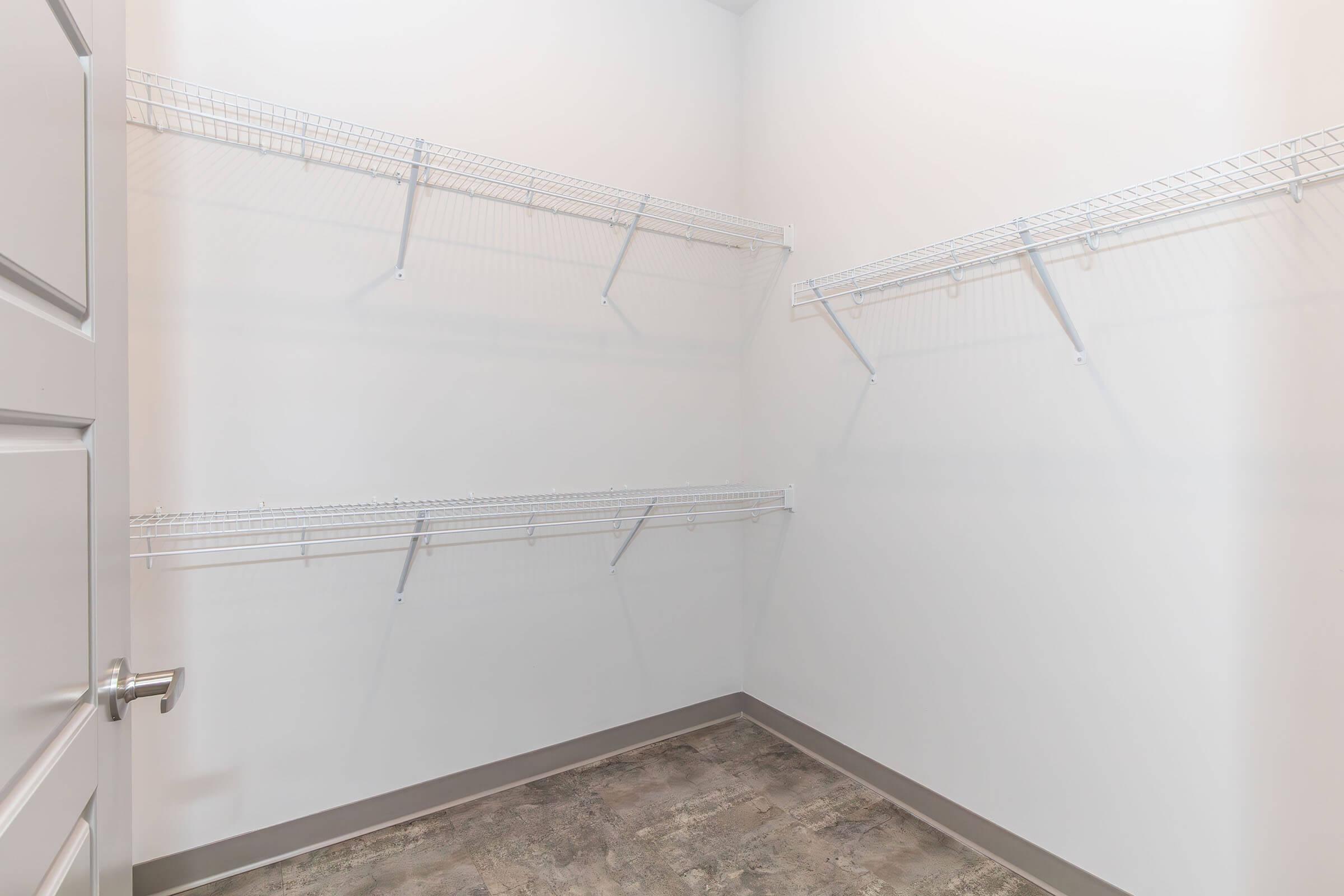 Empty closet with white walls and a light gray floor. Wire shelving is installed on two walls, with a door partially visible on the left. The space is bright and minimalist, providing a clean and organized look for storage.