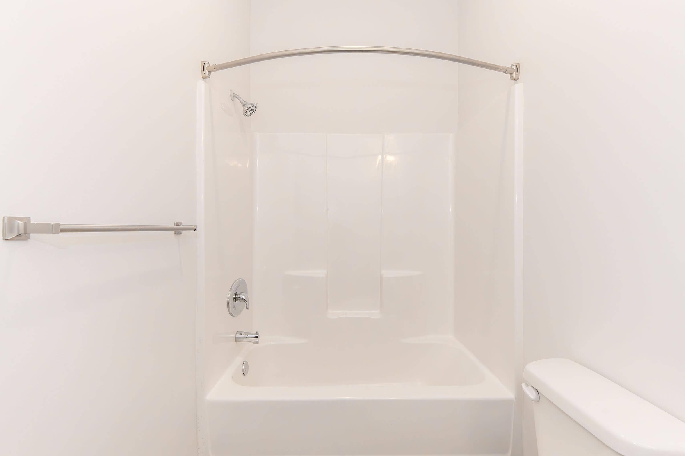 A clean, minimalist bathroom featuring a white bathtub with a curved shower rod and a showerhead. The walls are plain and white, enhancing the bright, airy feel of the space. A toilet is positioned to the right, and there's a towel bar on the wall. The overall design is simple and modern.