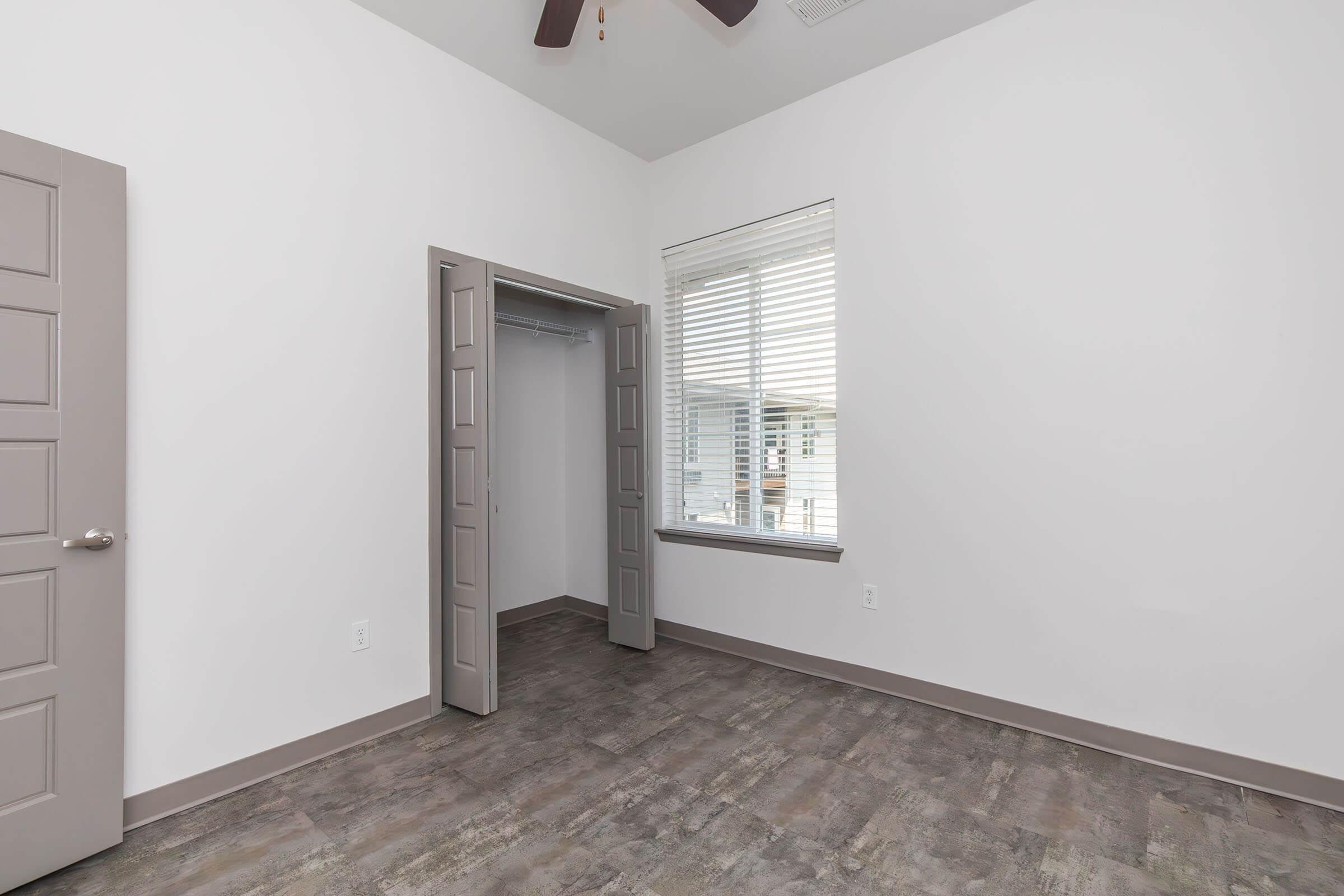 A small, empty room with white walls, a grey ceiling fan, and a large window featuring white blinds. There is a closet with double doors on one side and a grey floor that resembles wood. Natural light enters the room, giving it a bright and airy feel.