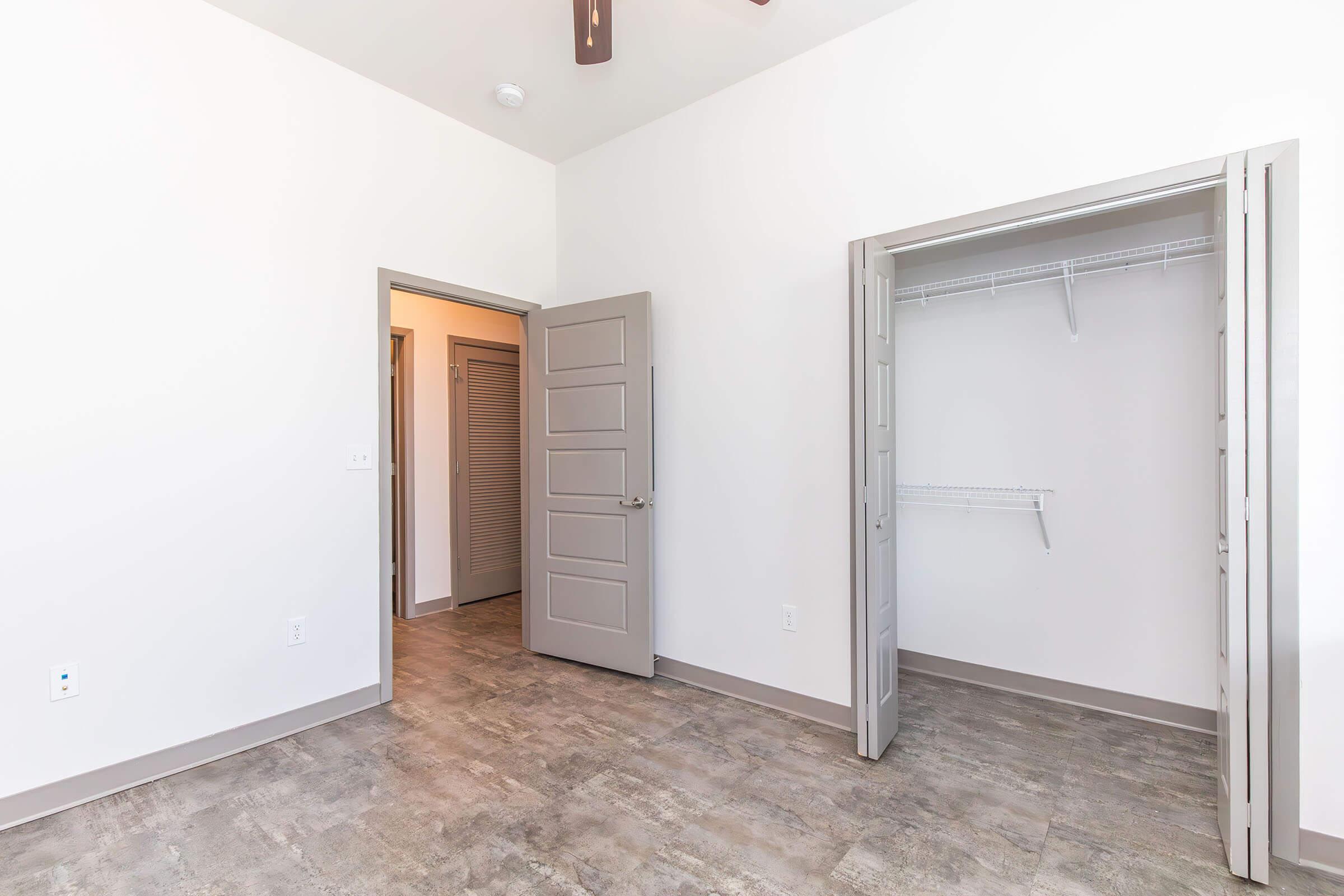A spacious, light-colored room with gray walls and a ceiling fan. Two doorways lead to adjacent areas; one door opens to a closet with shelving. The flooring is a modern, neutral tile, contributing to a fresh and airy atmosphere.