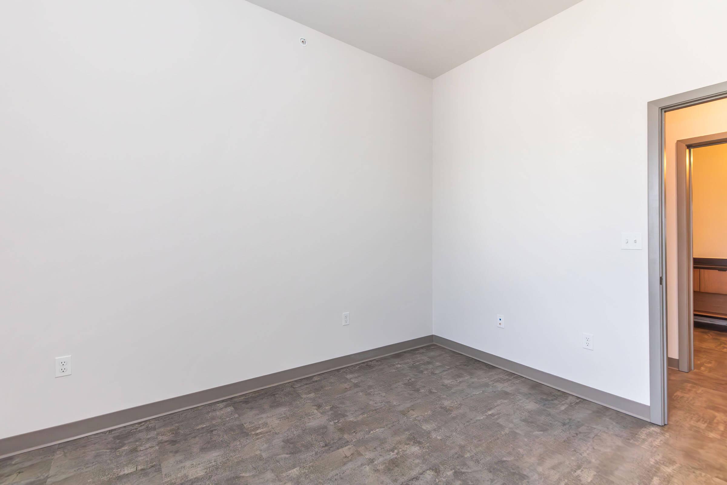 Empty room with light-colored walls and a gray floor. There is a doorway on the right leading to another space. The room appears clean and well-lit with natural light.