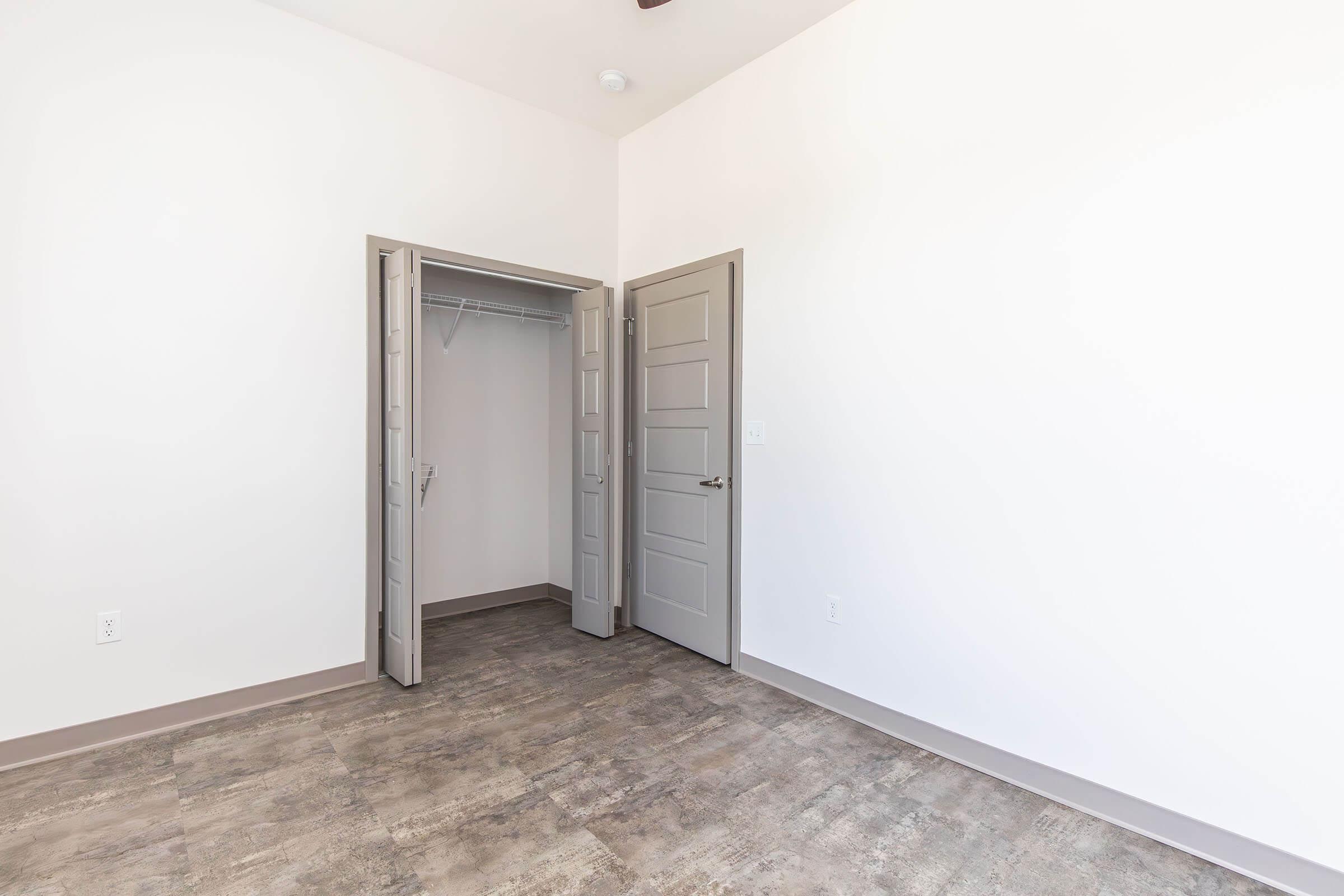A bright, empty room featuring light-colored walls and gray tile flooring. There are two closed doors: one leading to a closet on the left and another leading to an adjacent space on the right. The ceiling is white, and a light fixture is visible. The overall feel is modern and minimalistic.